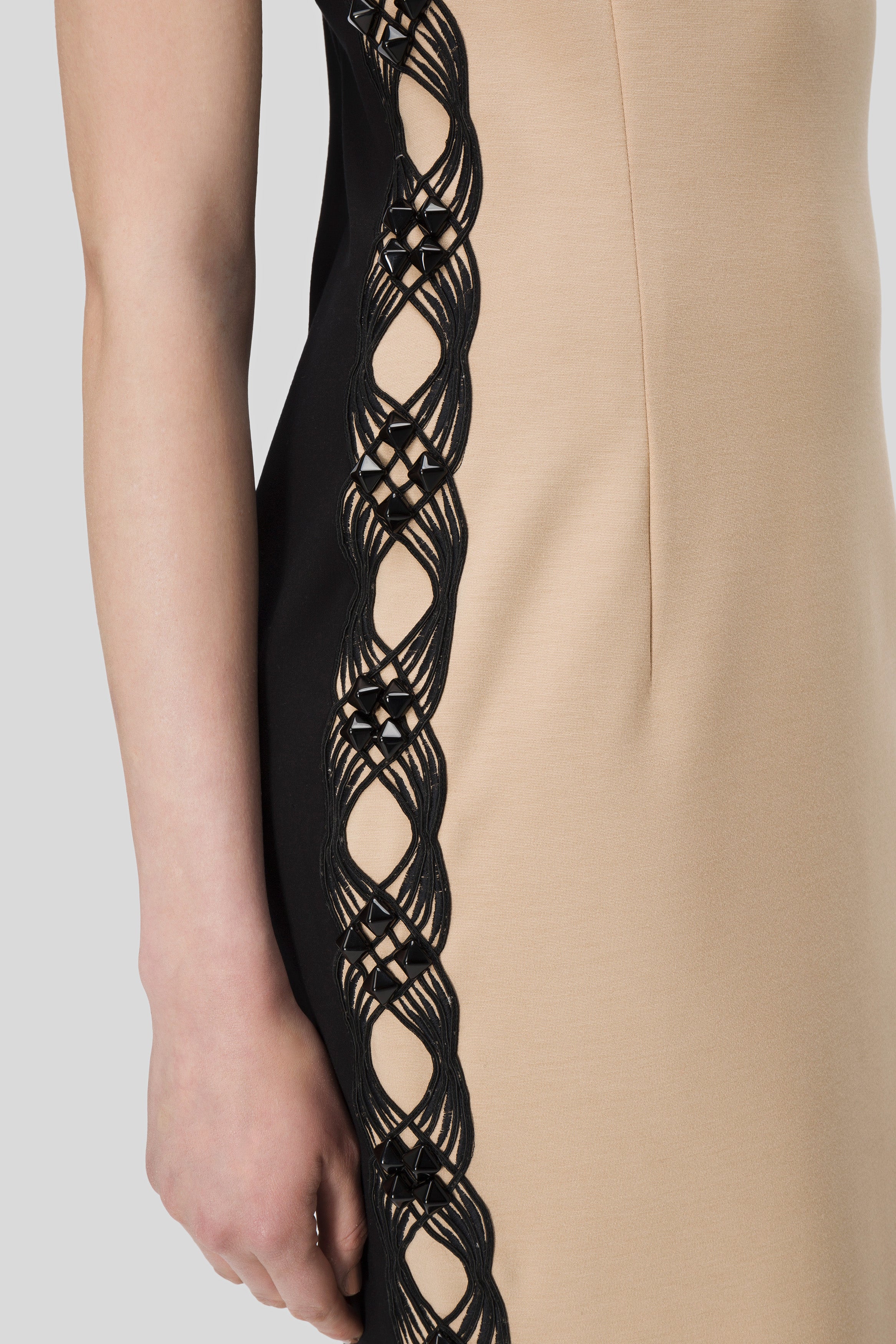 Close-up of an elegant Antonio Croce Adelin Sleeveless Dress featuring a beige front and a striking black side panel. The strapless neckline enhances its sophistication, with the panel showcasing intricate black lattice detailing that subtly reveals the skin beneath. You can see the arm of a person wearing this stunning dress by Antonio Croce.