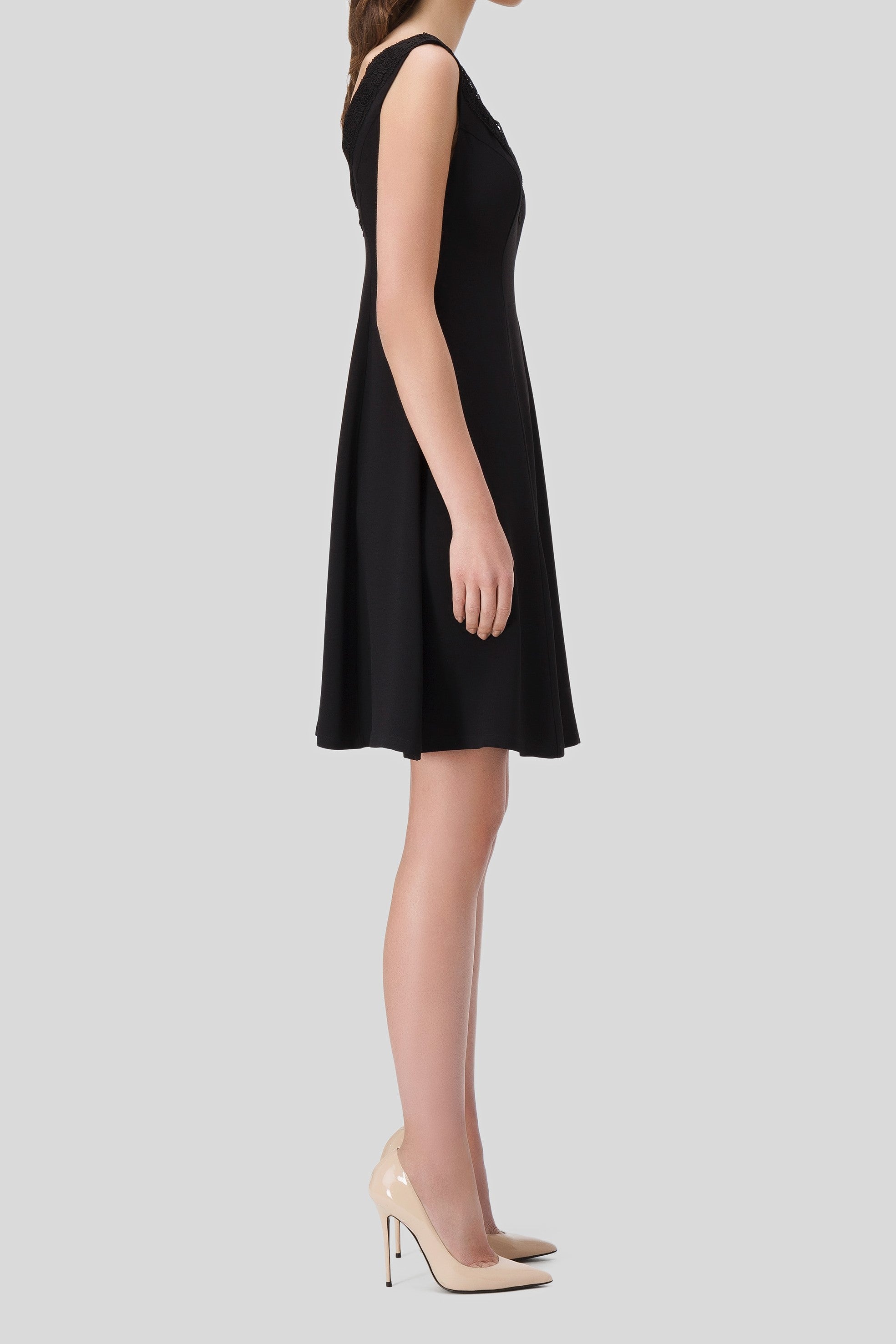 A person is standing in profile wearing the Antonio Croce Aira Romantic Black Dress, featuring an A-line silhouette and a slightly flared skirt that falls above the knees, paired with beige high heels against a plain grey background.