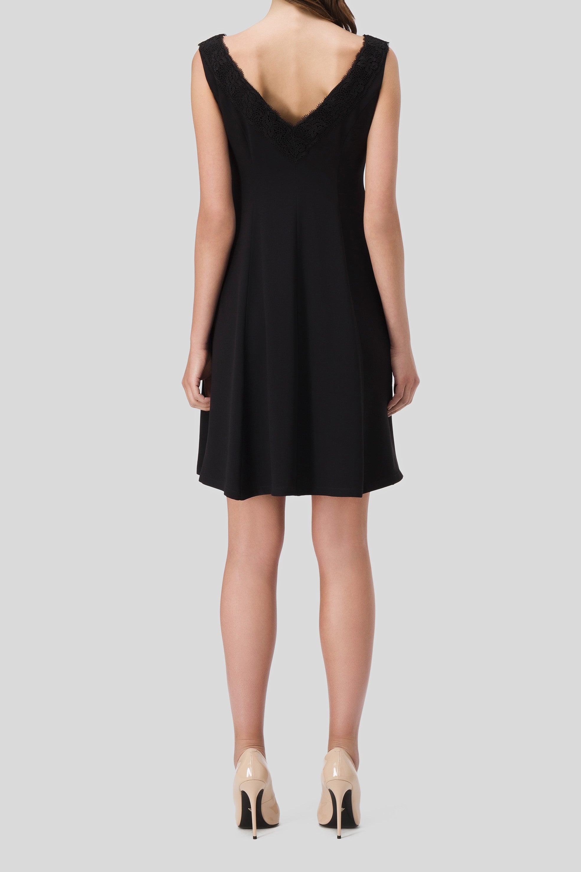 A woman is standing with her back to the camera, dressed in the Antonio Croce Aira Romantic Black Dress, which showcases an elegant A-line silhouette and a V-shaped neckline. She pairs it with nude high heels against a plain gray background.