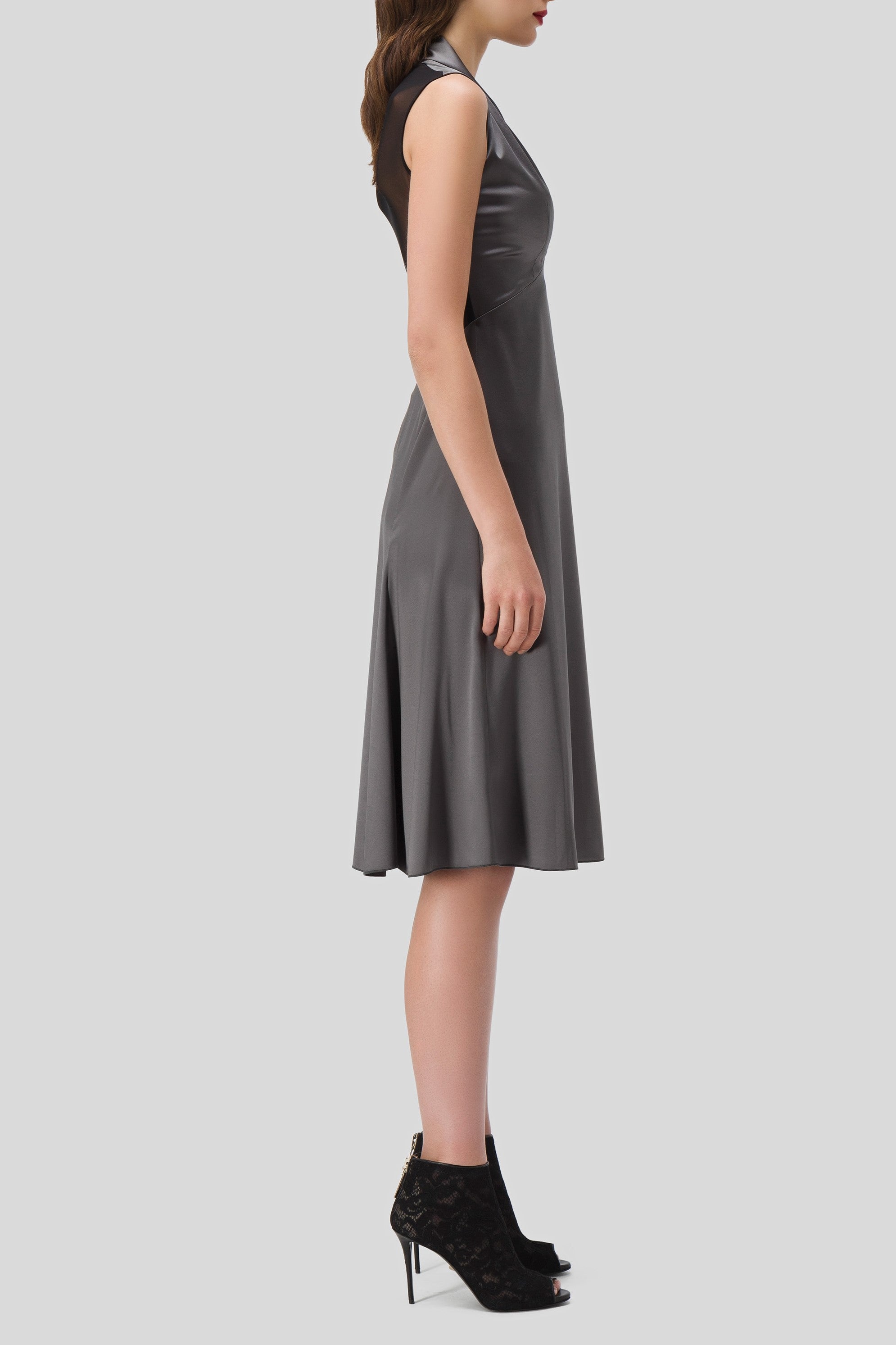 A person is wearing the Antonio Croce Akela Sleeveless Dress with Mesh Detail, a knee-length gray piece that embodies minimalist design, as they stand in profile against a light background. They complement the dress with black high-heeled ankle boots featuring lace details, echoing the essence of a vintage piece.
