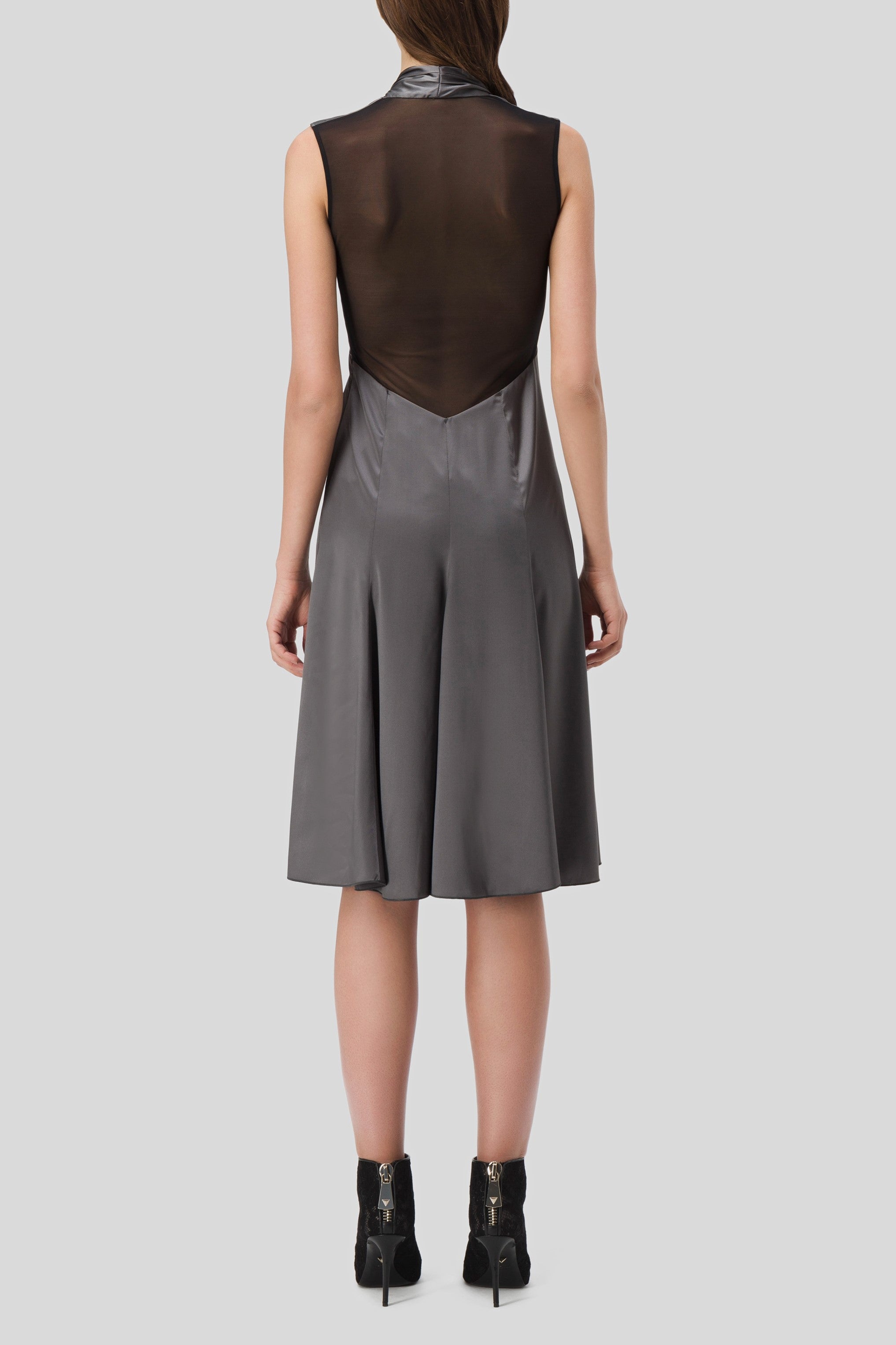 A person is wearing the Antonio Croce Akela sleeveless dress with mesh detail, showcasing a knee-length gray design with a sheer black back panel that exudes minimalism against a plain backdrop. The vintage-inspired piece features a flared skirt and is perfectly paired with black ankle boots featuring zippers.