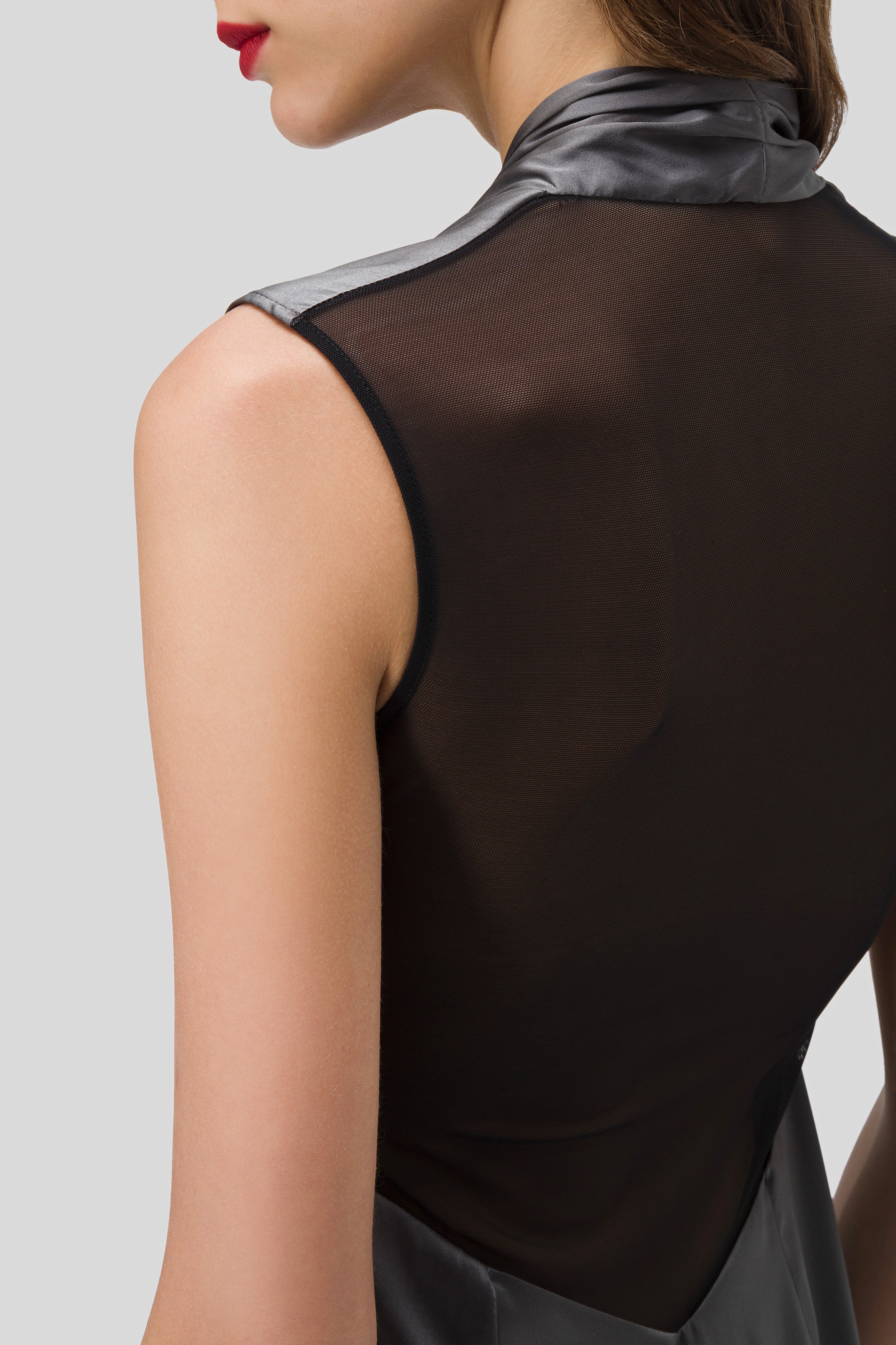 A woman is wearing an Antonio Croce Akela Sleeveless Dress featuring a sheer mesh detail on the back and a minimalist shiny silver collar. Her hair is tucked behind her ear, and she has bold red lipstick. The background is plain gray.
