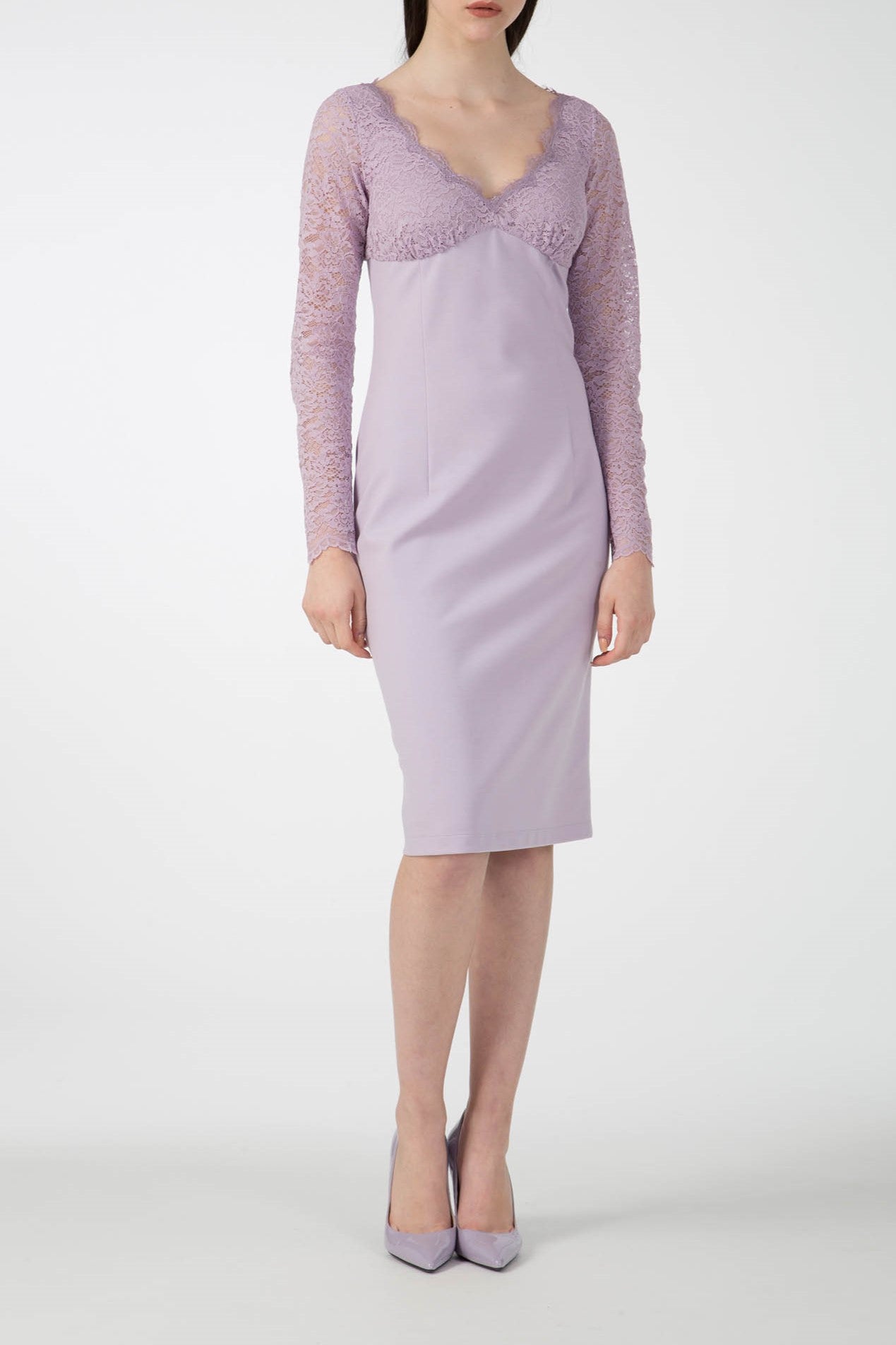 A woman is wearing the form-fitting Antonio Croce Ampara Embroidered Lace Dress, distinguished by its long sleeves and scalloped neckline crafted from delicate lace fabric. She pairs it elegantly with matching purple heels as she stands against a plain white background.