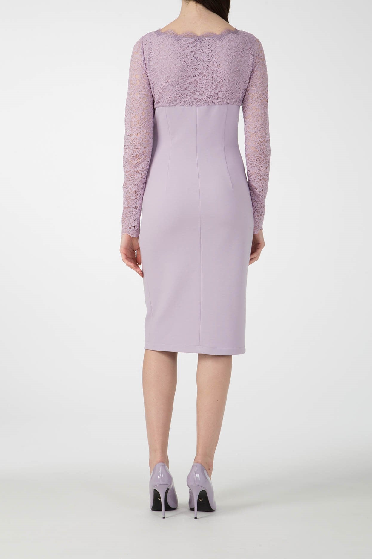 A person wearing the Antonio Croce Ampara Embroidered Lace Dress, an elegant lavender knee-length outfit with delicate lace sleeves, and matching high heels stands against a plain white background, facing away from the camera.