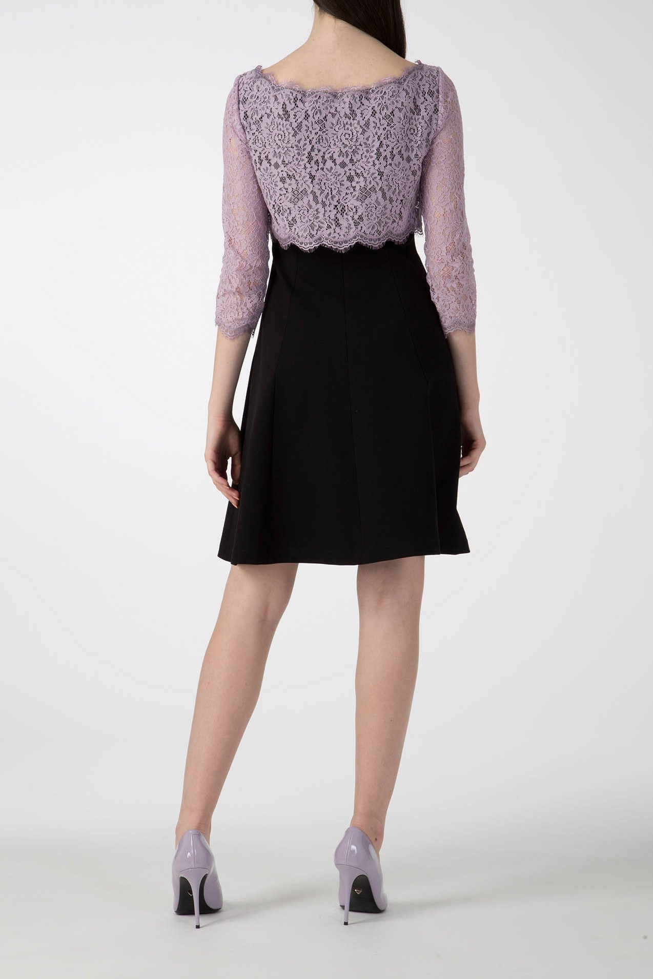 A woman in the sophisticated Antonio Croce Artemide Two-Tone Dress, featuring an elegant A-line silhouette with a black skirt and a light purple lace bodice and three-quarter sleeves, stands with her back to the camera. Her long hair falls straight, complementing her light purple high-heeled shoes.