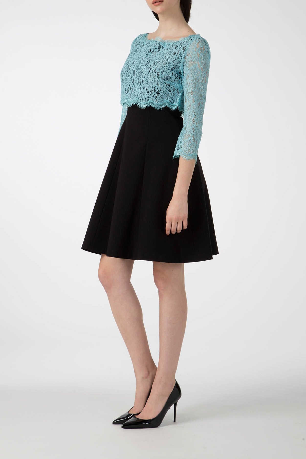 A woman is wearing the Antonio Croce Artemide Two-Tone Dress, featuring a teal lace bodice with three-quarter sleeves and a black A-line silhouette skirt. She is standing sideways in black high heels against a plain white background.