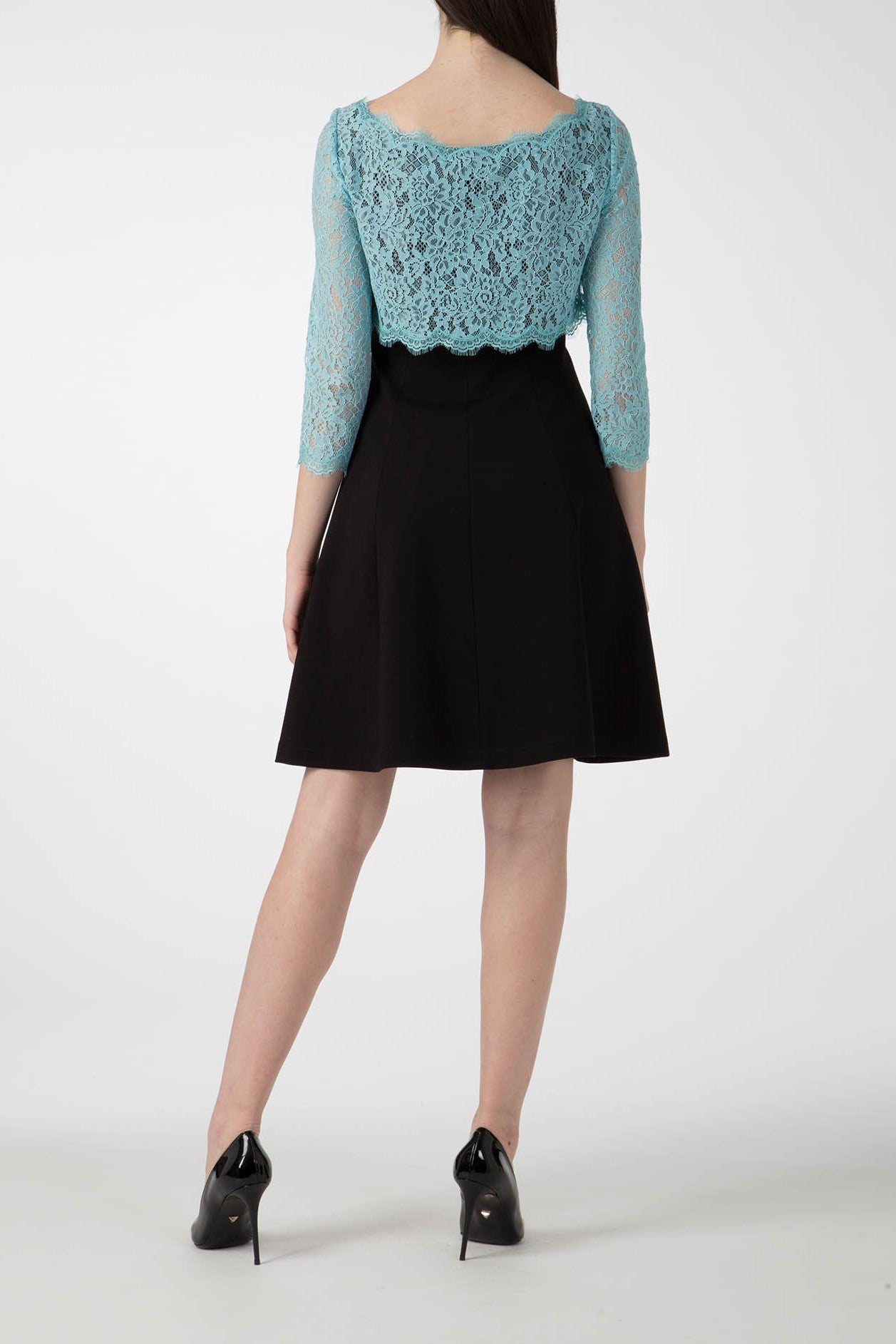 A person stands facing away, elegantly dressed in the Antonio Croce Artemide Two-Tone Dress, which features a blue lace bodice with three-quarter sleeves and a black A-line silhouette skirt. They complete the look with black high heels against a plain white background.