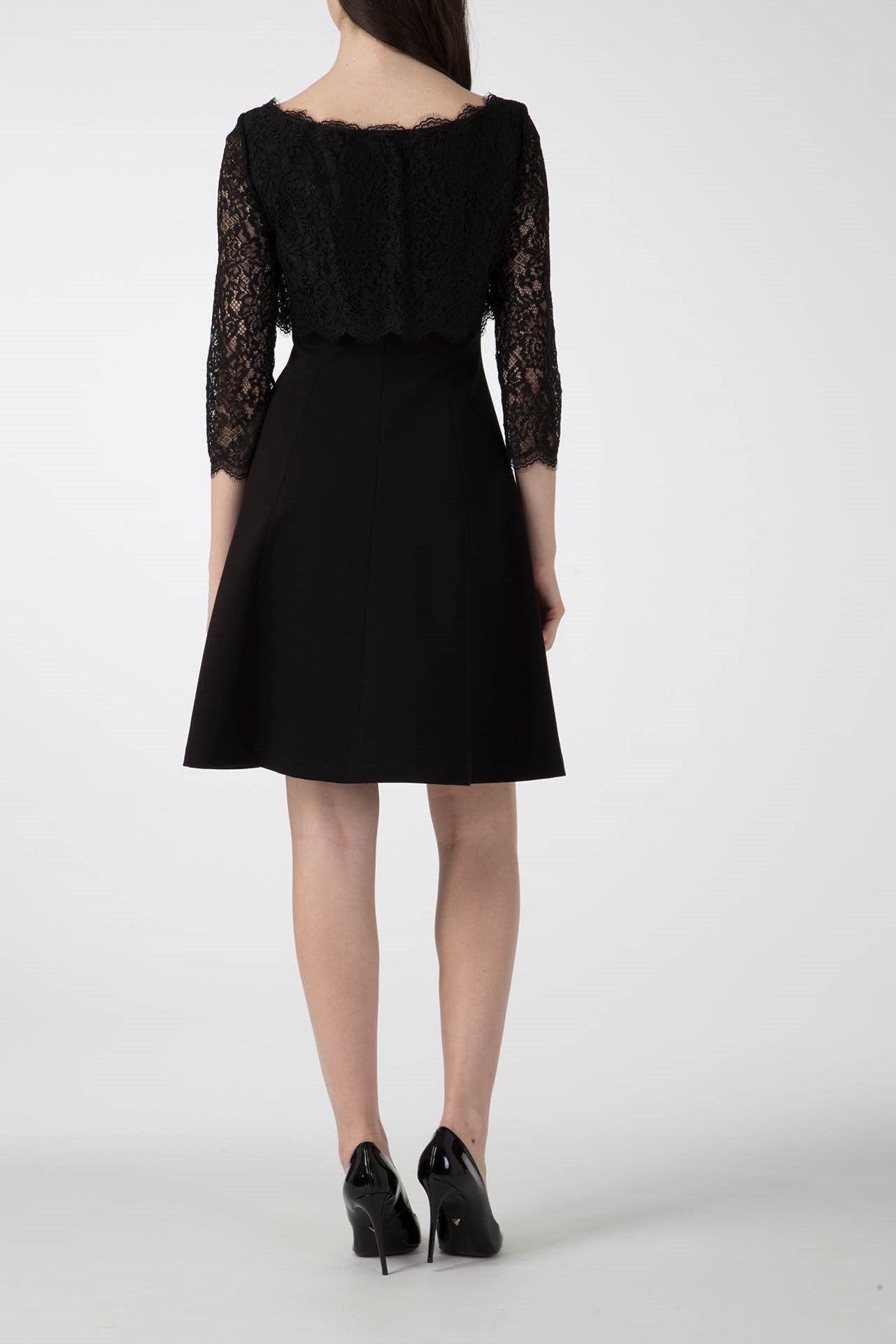 A woman in an elegant Antonio Croce Artemide Two-Tone Dress stands facing away. The knee-length A-line silhouette, complemented by three-quarter sleeves and an intricate lace bodice, enhances her allure. Paired perfectly with black high heels, the plain light-colored background highlights her sophisticated style.