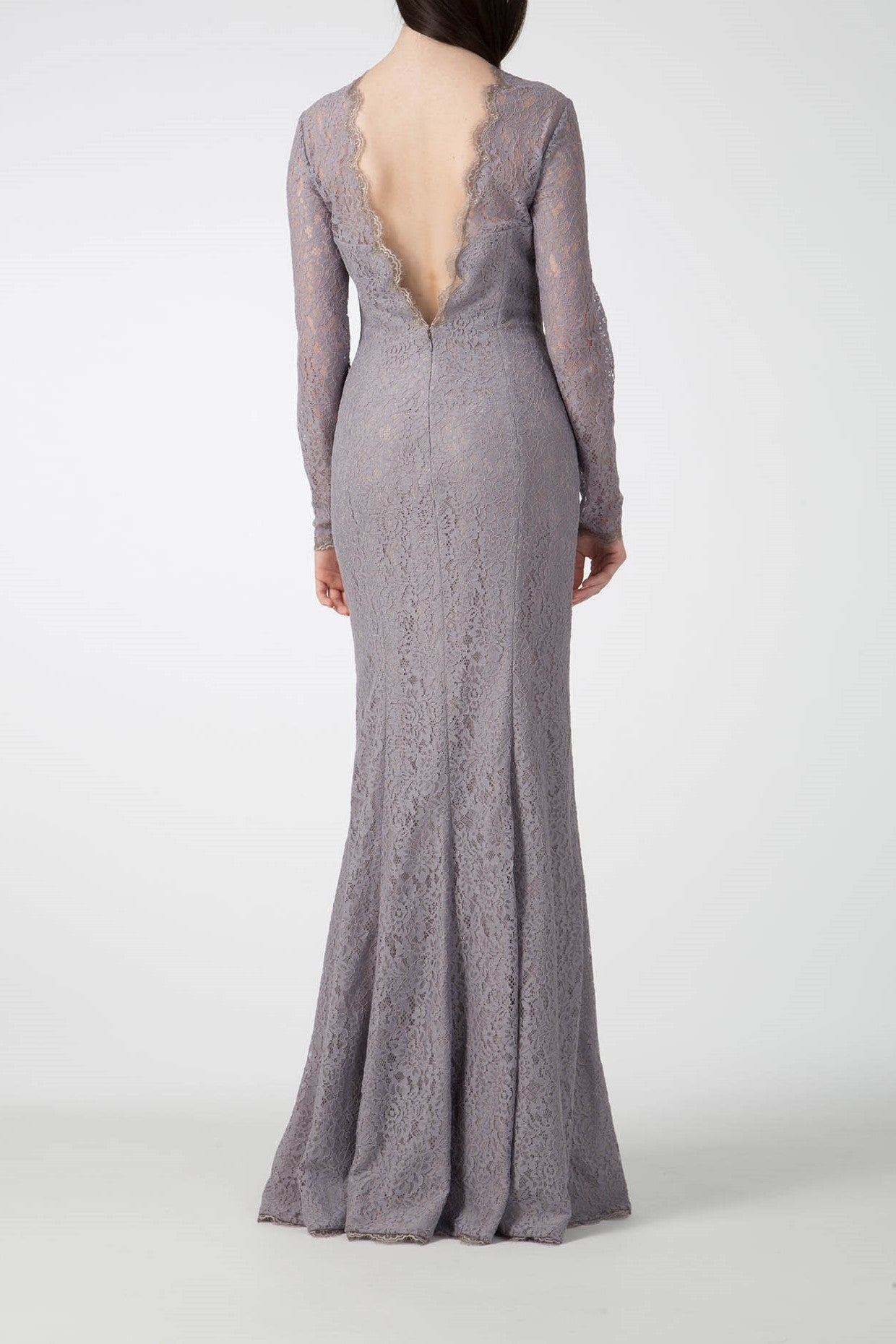 A woman stands facing away, elegantly attired in the Antonio Croce Australia Allover Lace Gown with Plunging Back. Her form-fitting, floor-length lavender dress, adorned with intricate lace and a deep V-back, beautifully complements her long, dark hair cascading gracefully down her back against the plain white background.