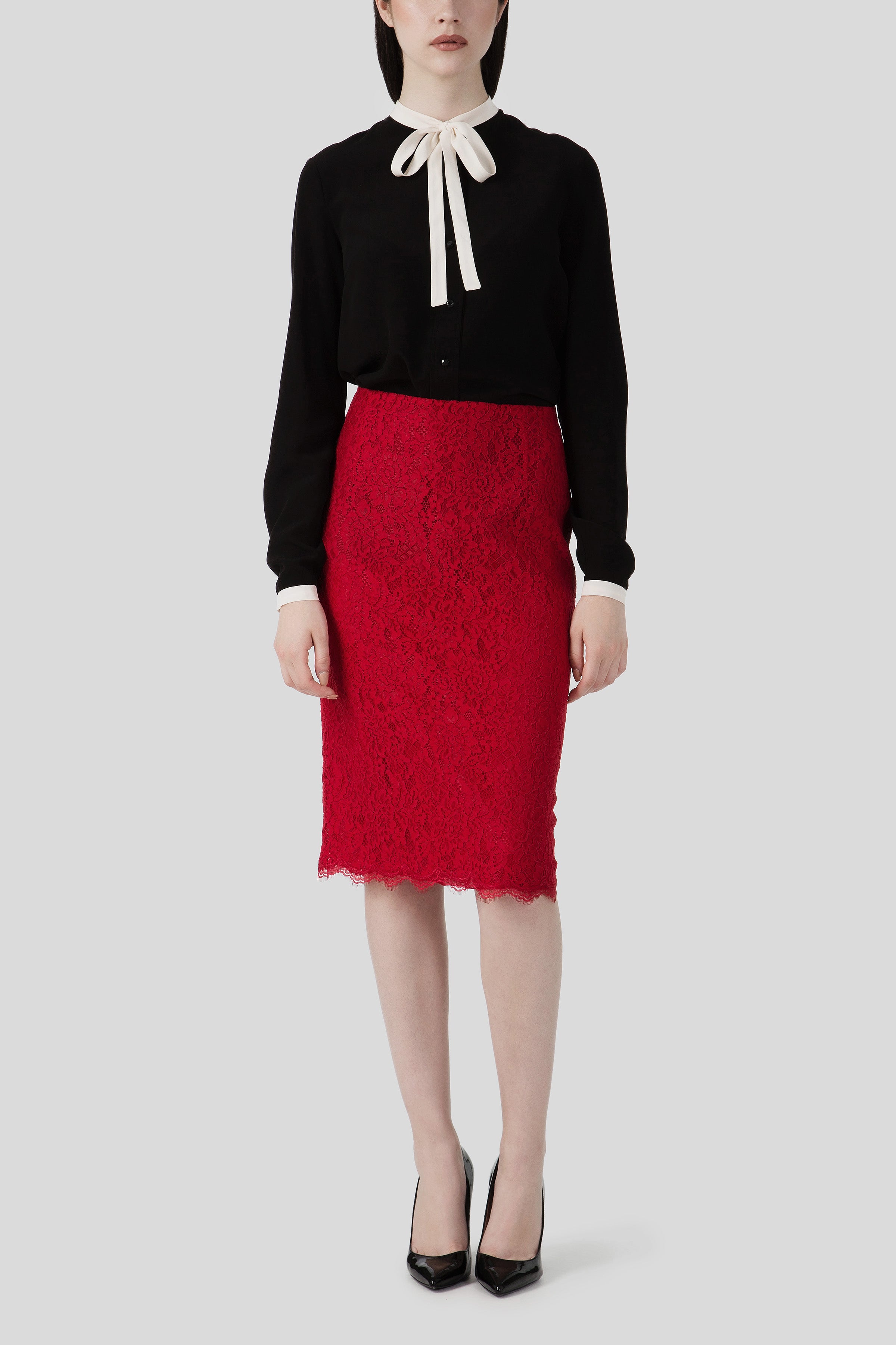 Person wearing a black blouse with a white bow tie and a romantic Antonio Croce Scarlett Lace Skirt in red, paired with black heels, standing against a plain background.