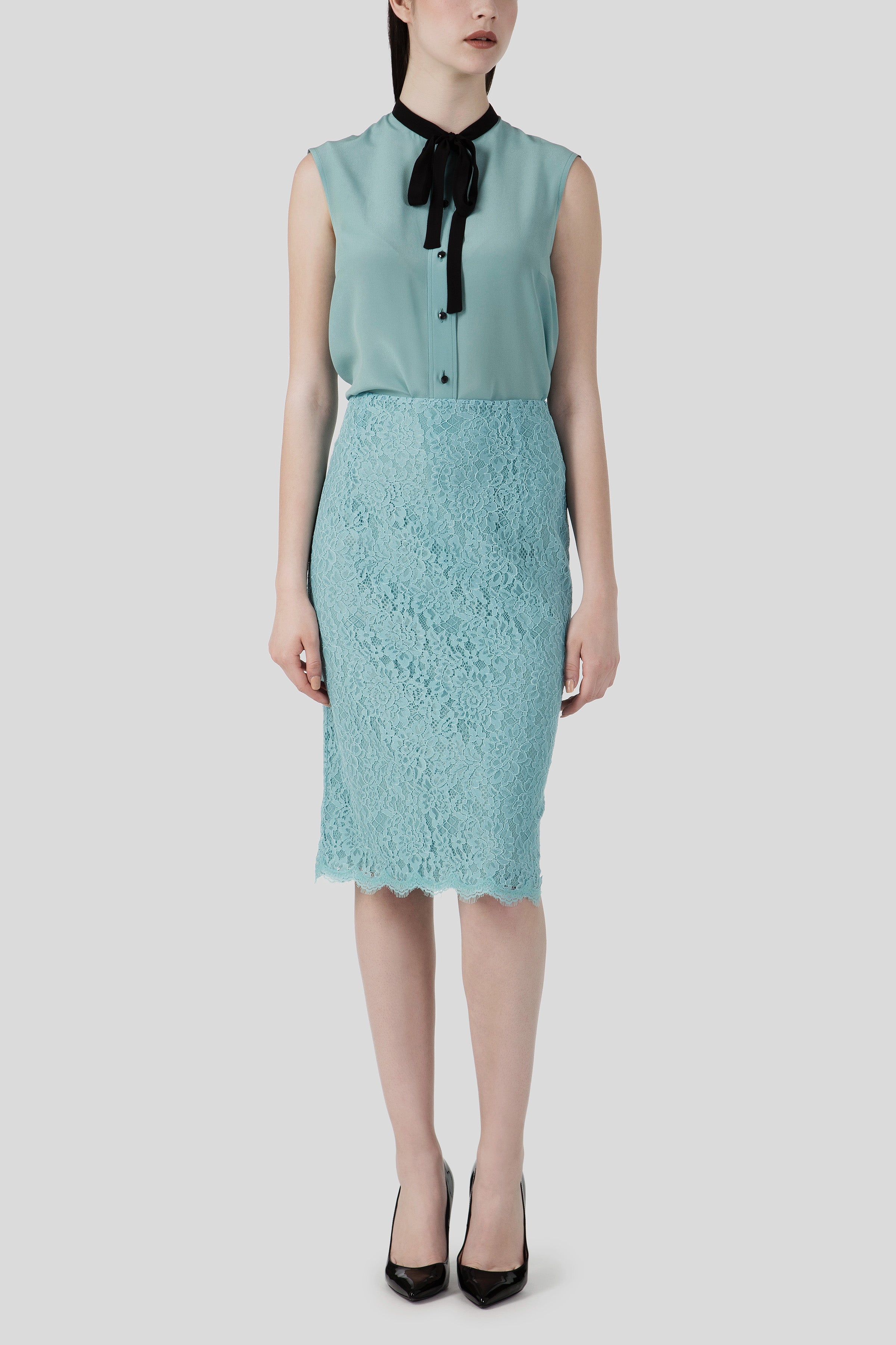 The woman is dressed in an Antonio Croce Bibi flowy blouse with a tie neck, paired with a vintage lace pencil skirt and a black necktie. Against a plain grey backdrop, her outfit is completed with black high-heeled shoes.