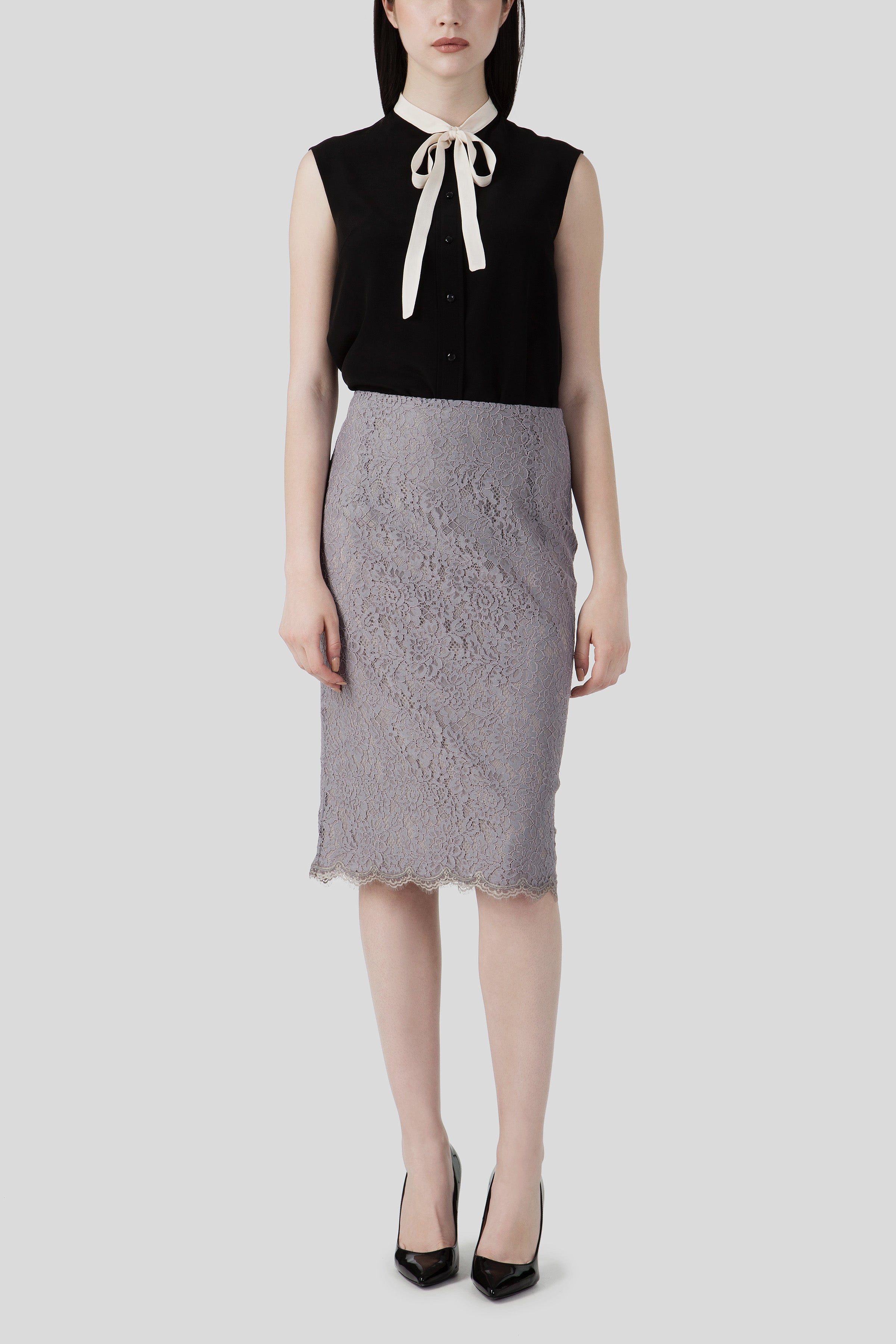 A woman stands gracefully against a plain backdrop, wearing the vintage Antonio Croce Bibi Flowy Blouse with Tie Neck, artfully paired with a knee-length gray lace skirt. Her ensemble is completed by elegant black high heels, adding sophistication to her classic look.