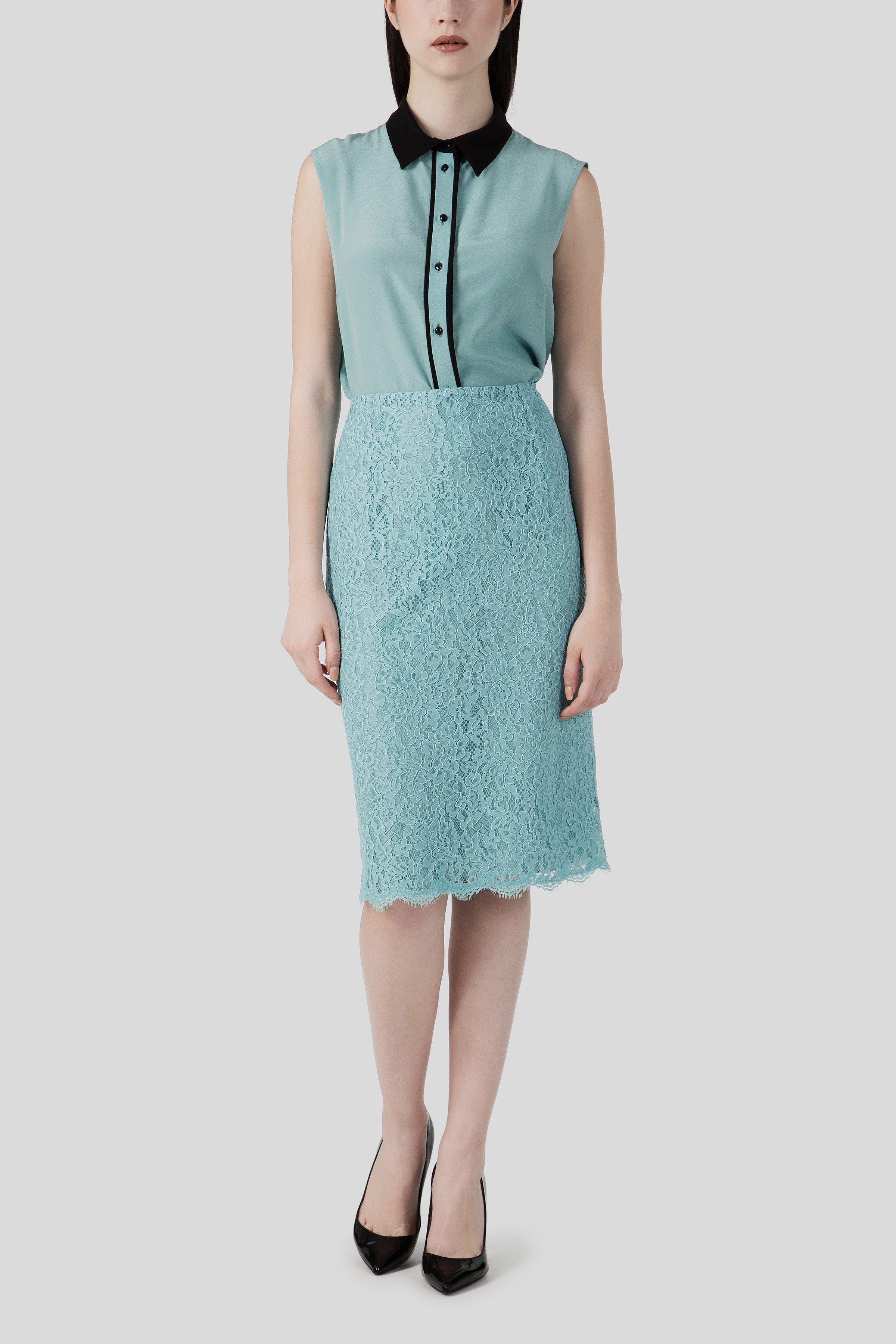 A person is wearing a sleeveless teal blouse with a black collar and buttons, paired with the Antonio Croce Scarlett Lace Skirt in the same shade. They are standing against a plain gray background and wearing black pointed heels, showcasing an effortlessly chic style.