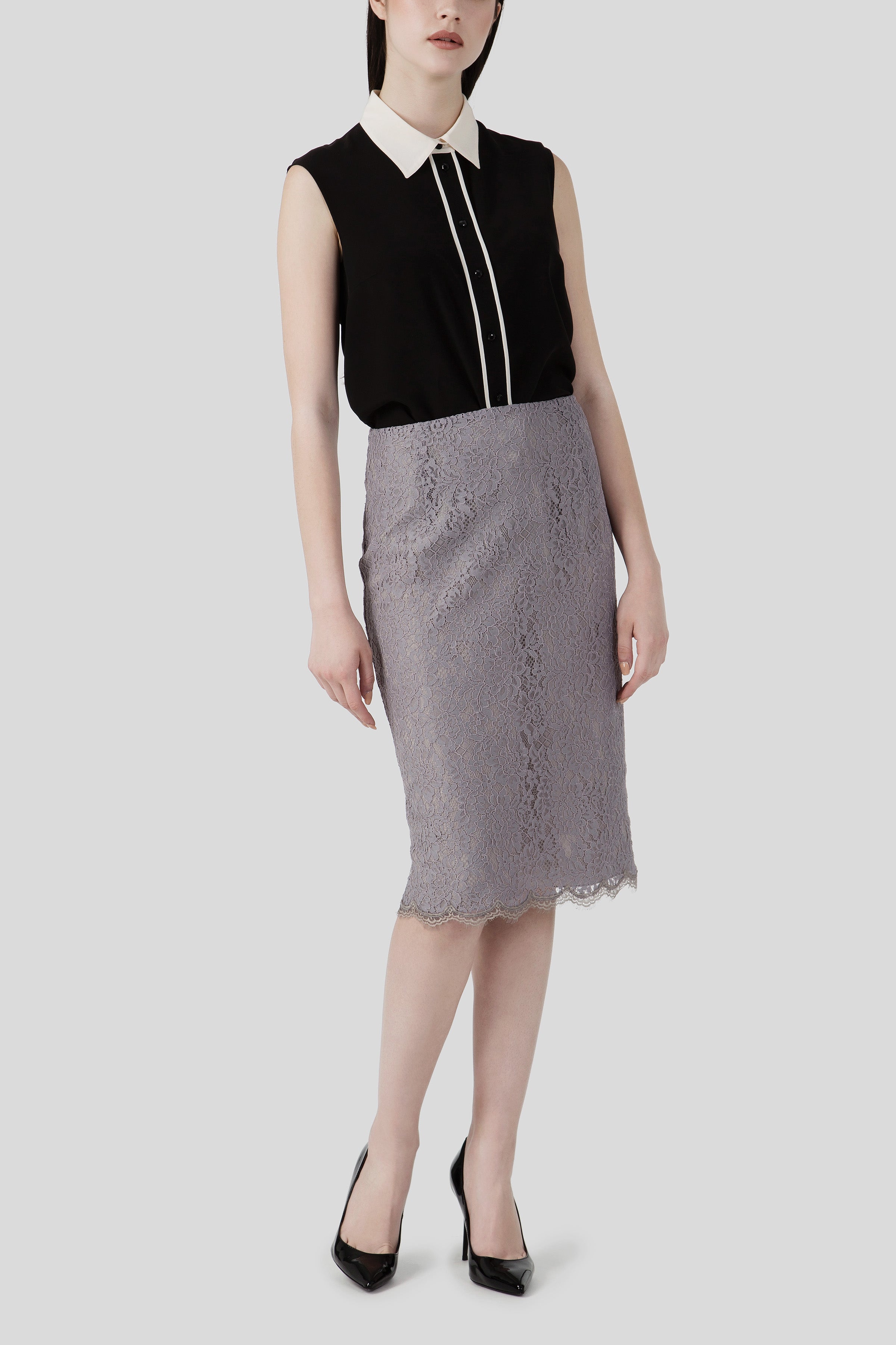 A person is elegantly dressed in a vintage piece: the Antonio Croce Brandy Sleeveless Shirt, featuring a contrasting white collar, paired with a knee-length grey lace skirt. They complete the look with black high-heeled shoes against a plain white background.