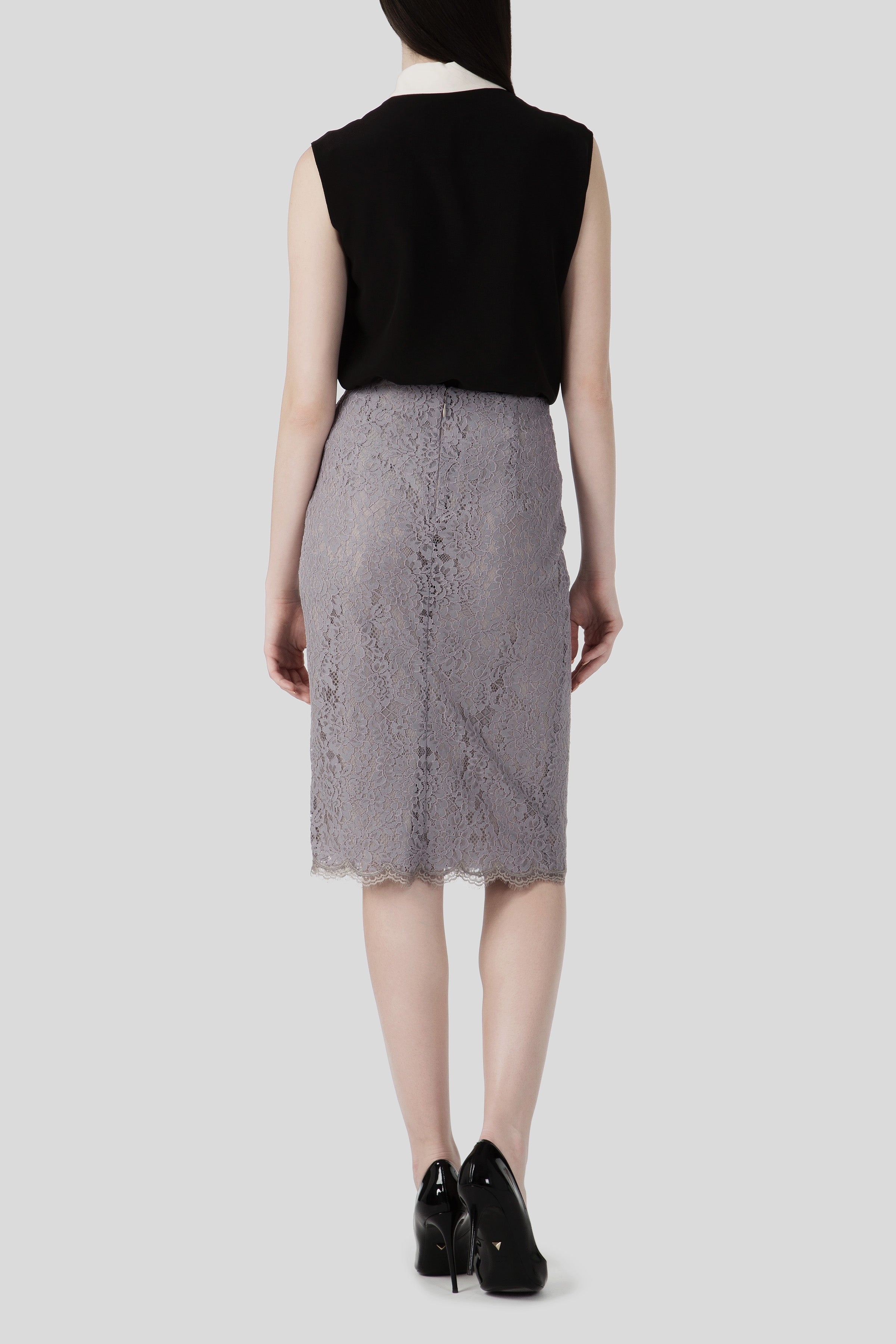 A woman stands with her back to the camera, elegantly dressed in a black sleeveless top paired with the Antonio Croce Scarlett Lace Skirt, which is knee-length and vintage in style. Her chic ensemble is completed with black high-heeled shoes, set against a plain light gray background.
