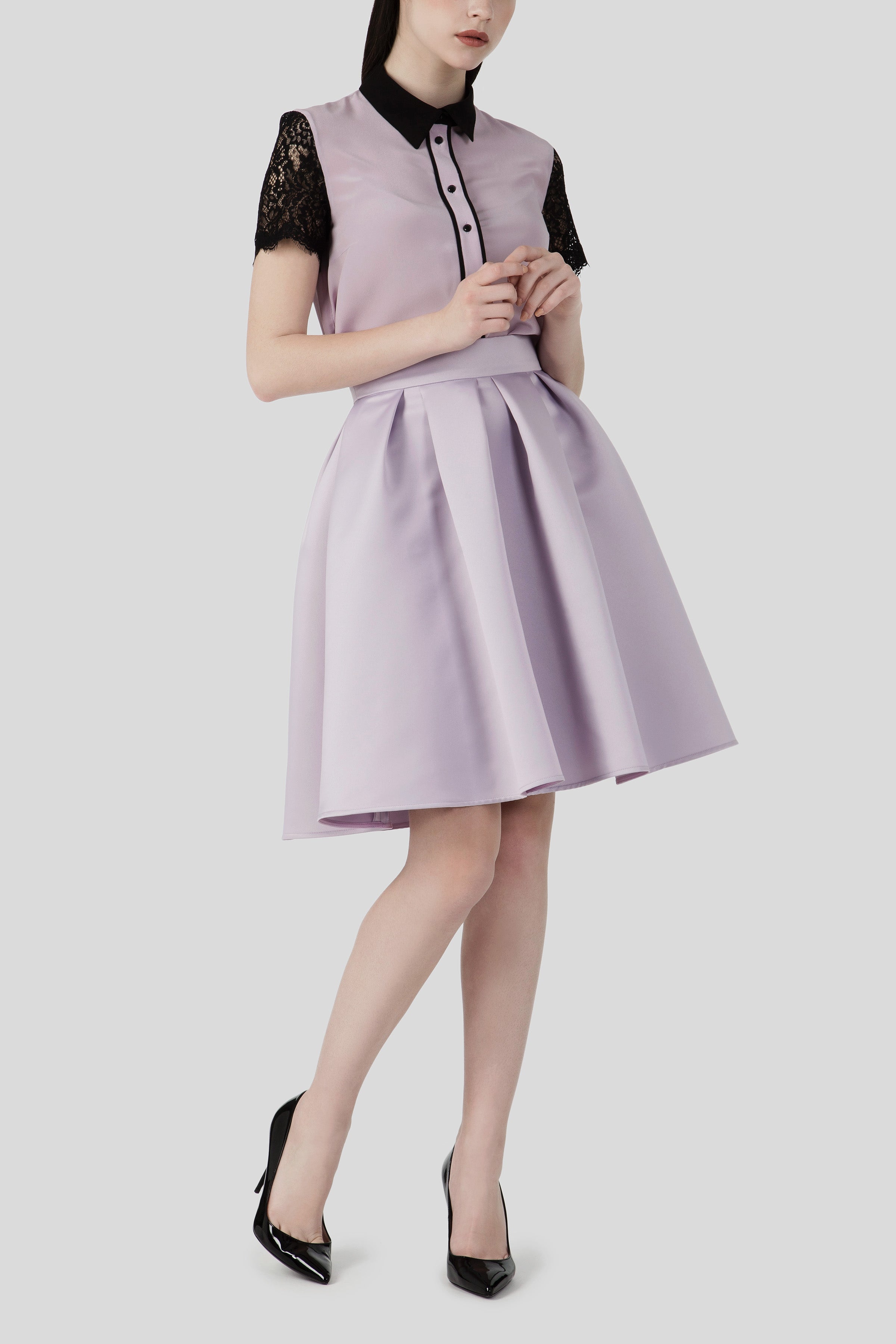 Against a gray background, a woman showcases an Antonio Croce ensemble featuring the Shelby A-Line Skirt, characterized by a flattering pleated silhouette. Her lavender dress is accentuated with black lace sleeves and collar, and she completes her stylish vintage look with black high-heeled shoes.