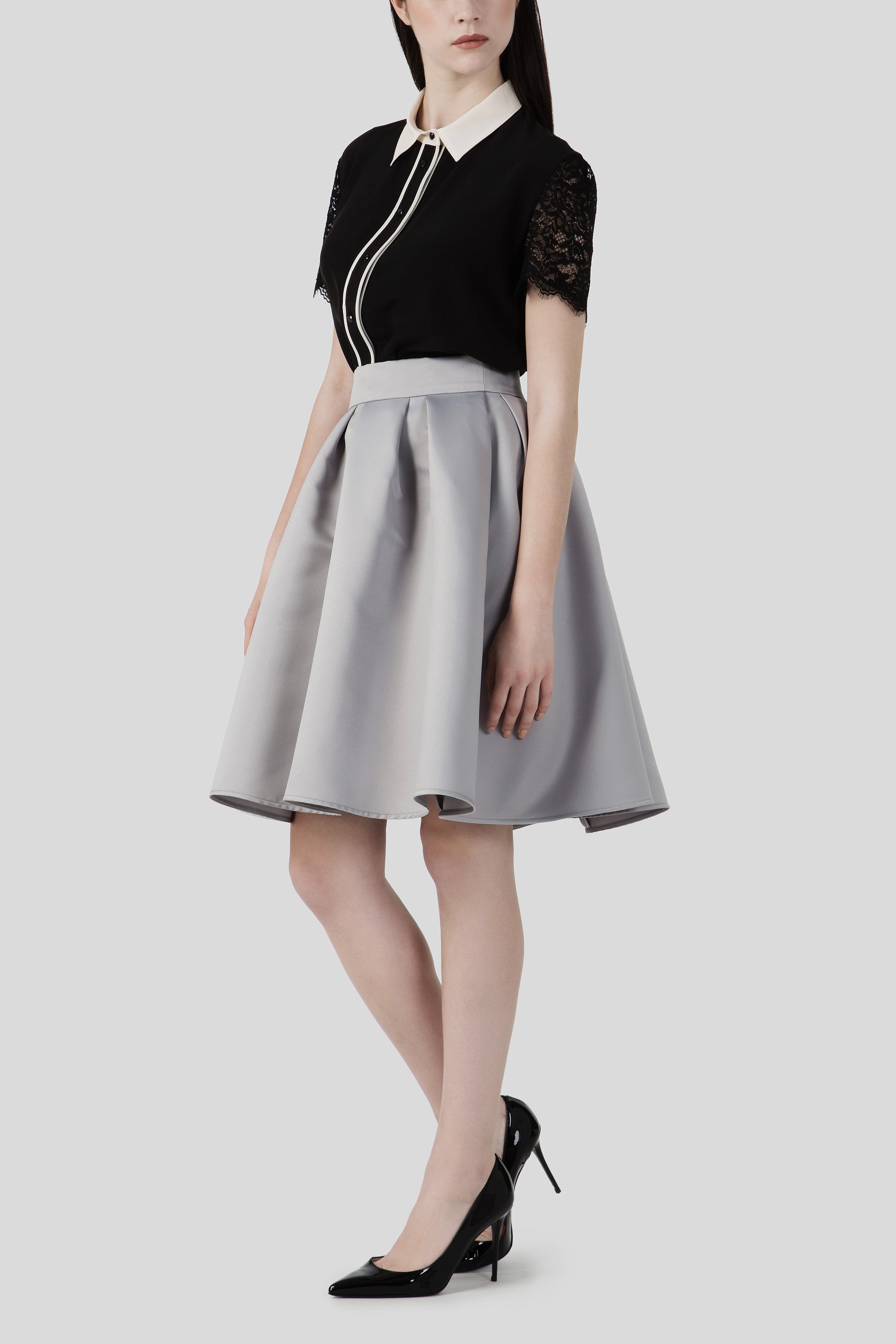 A woman is wearing a black blouse with lace sleeves and a white collar, paired with the Antonio Croce Shelby A-Line Skirt, which is vintage high-waisted gray and creates a flattering silhouette. She has on black high-heeled shoes against a plain light gray background.