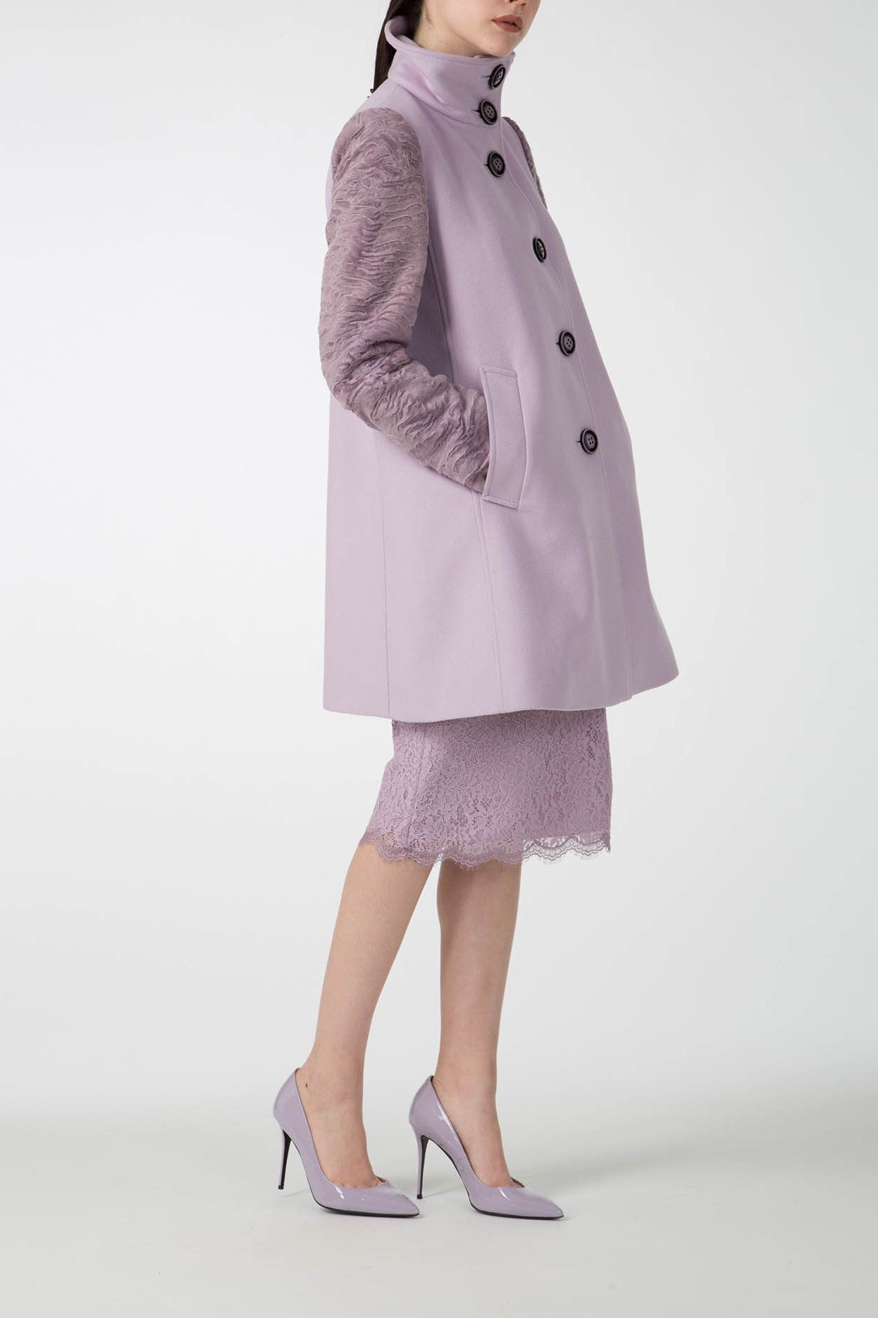 A person in profile showcases the Antonio Croce Callie Textured Coat with Contrast Sleeves, exuding style in lavender with its distinctive sleeve design. They complement the coat with a coordinated lace skirt and pointed high heels, set against a simple, light gray backdrop that accentuates the outfit.