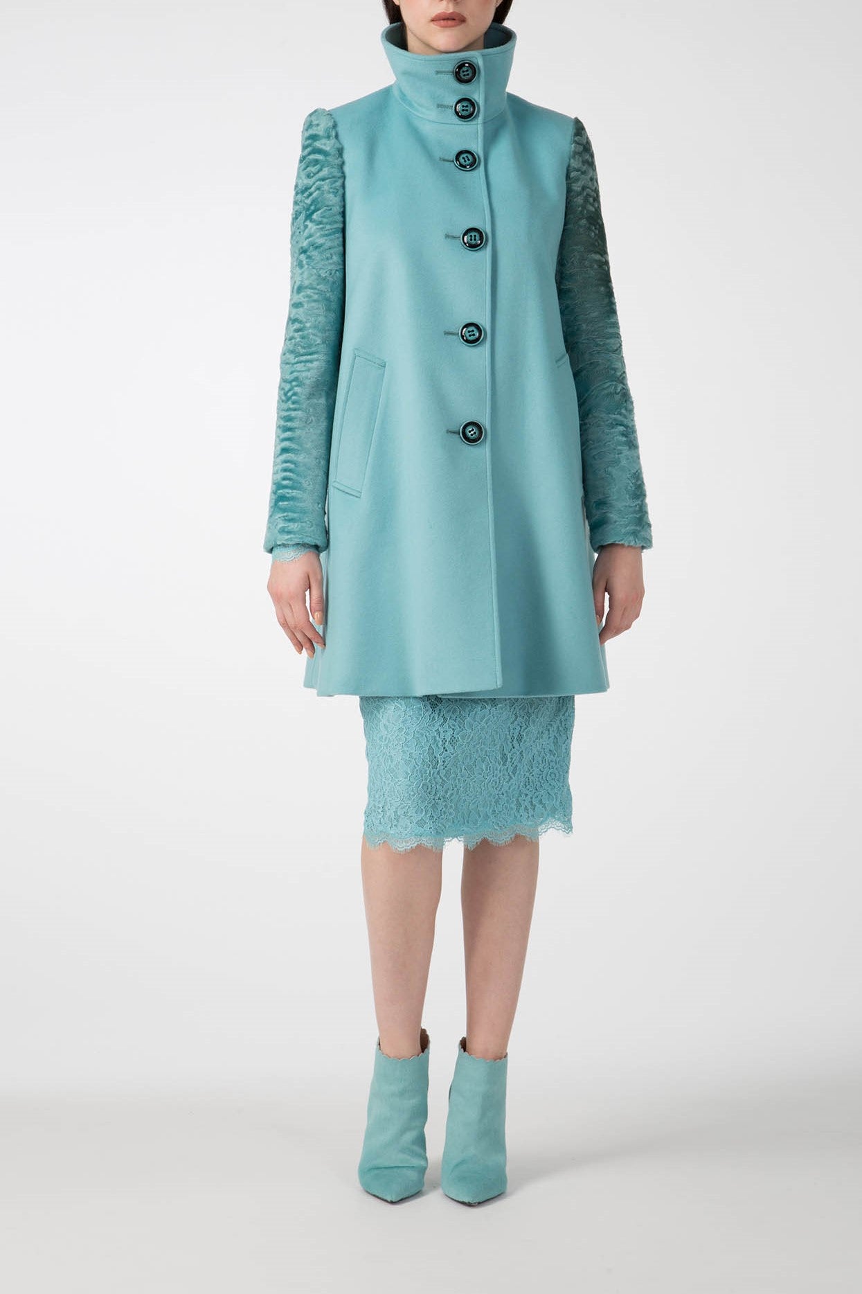 A person wearing the Antonio Croce Callie Textured Coat with Contrast Sleeves, a true vintage piece, stands facing forward. The stylish coat in teal features black buttons and unique sleeve details. Underneath is a matching teal lace skirt, complemented by teal ankle boots against a plain white background.