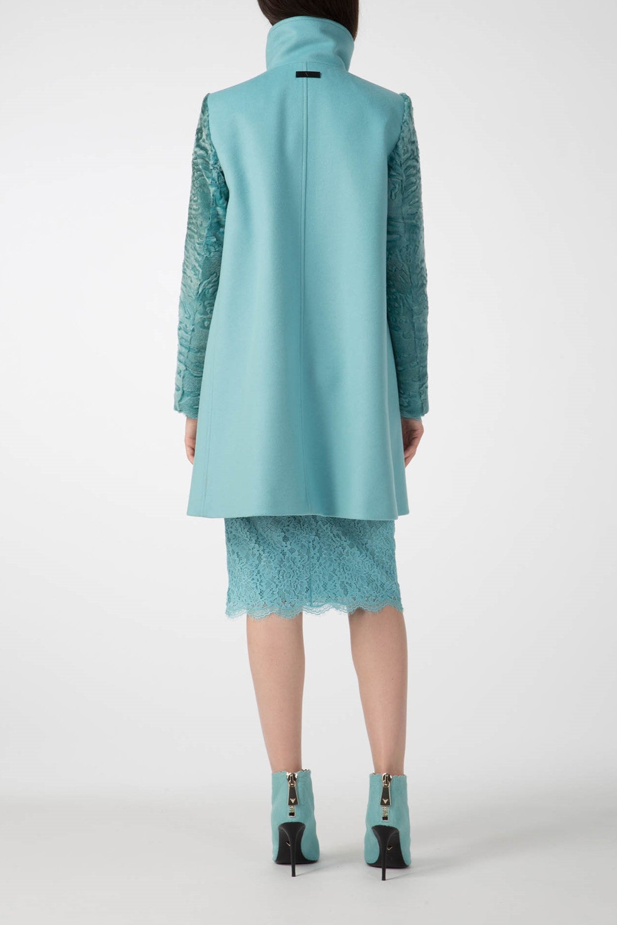 A person is wearing the Antonio Croce Callie Textured Coat with Contrast Sleeves, a stylish teal long coat featuring unique sleeve details, along with a matching lace dress. They also sport teal ankle boots with high heels as they stand with their back to the camera, set against a plain white background that highlights this vintage piece.