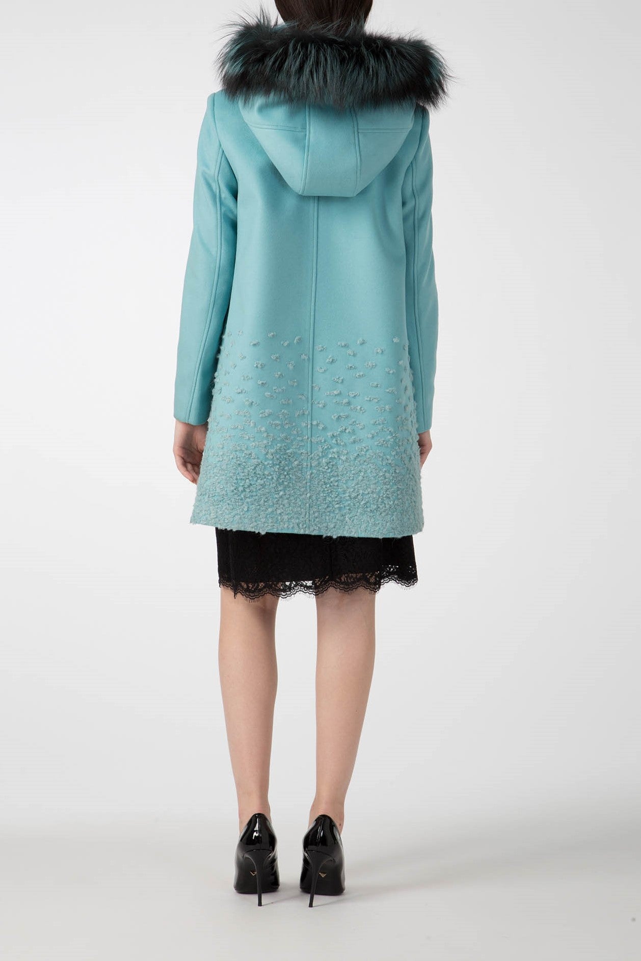 Dressed in the Antonio Croce Carolina Textured Coat with Hood, featuring a detachable fur hood, an individual stands with their back to us, showcasing the distinct texture on the lower portion of the coat. They combine it with a black lace skirt and glossy black shoes to create an elegant winter look against a simple white backdrop.