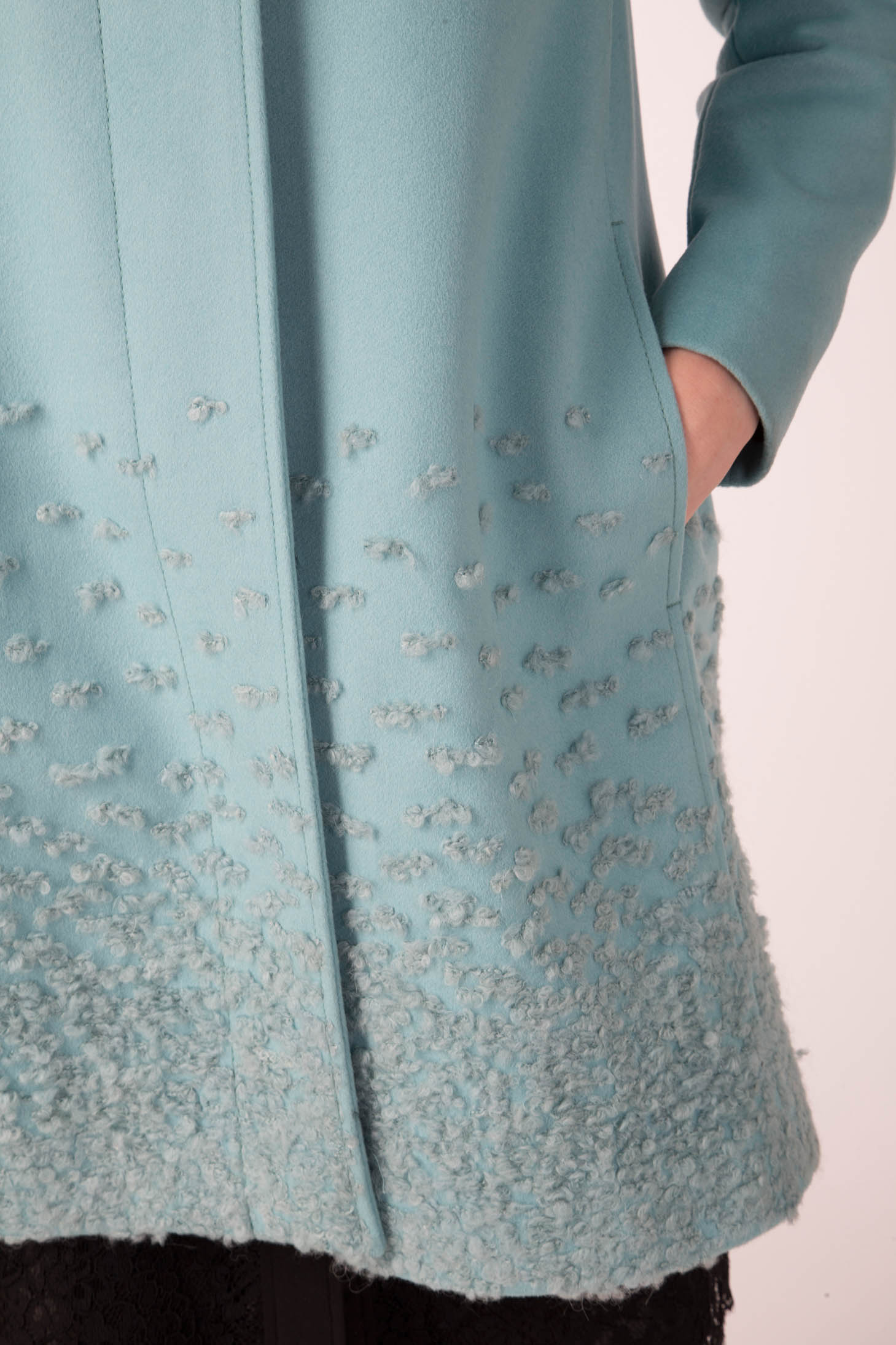 Close-up of a person wearing the Antonio Croce Carolina Textured Coat with Hood in light teal, showcasing fabric details at the hem. The coat, from the brand Antonio Croce, features a pocket where the person's hand is partially visible and includes a detachable fur hood, all set against a plain background.