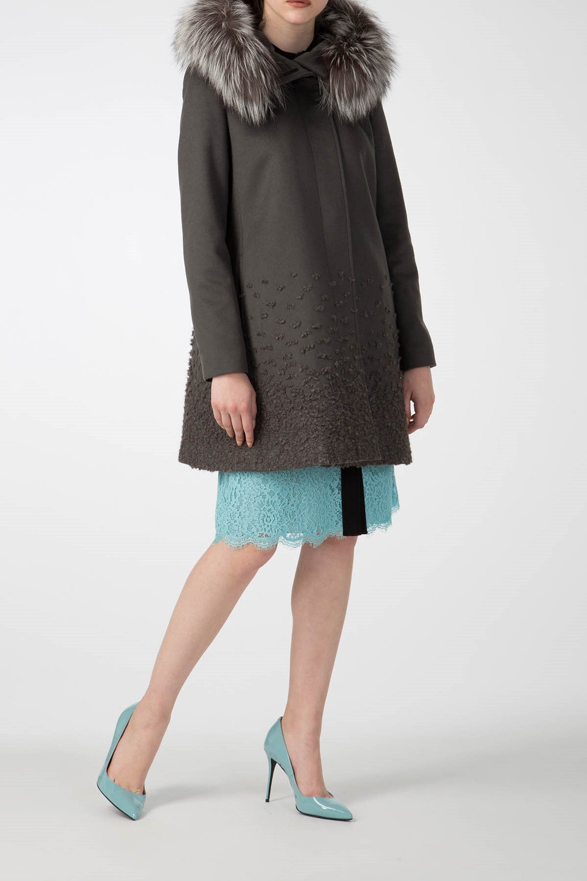 The Antonio Croce Carolina Textured Coat with Hood is showcased in a stylish winter ensemble. This chic dark overcoat, complete with a detachable fur hood and textured lower fabric, beautifully complements a turquoise lace skirt and matching high heels against a minimalist white background.
