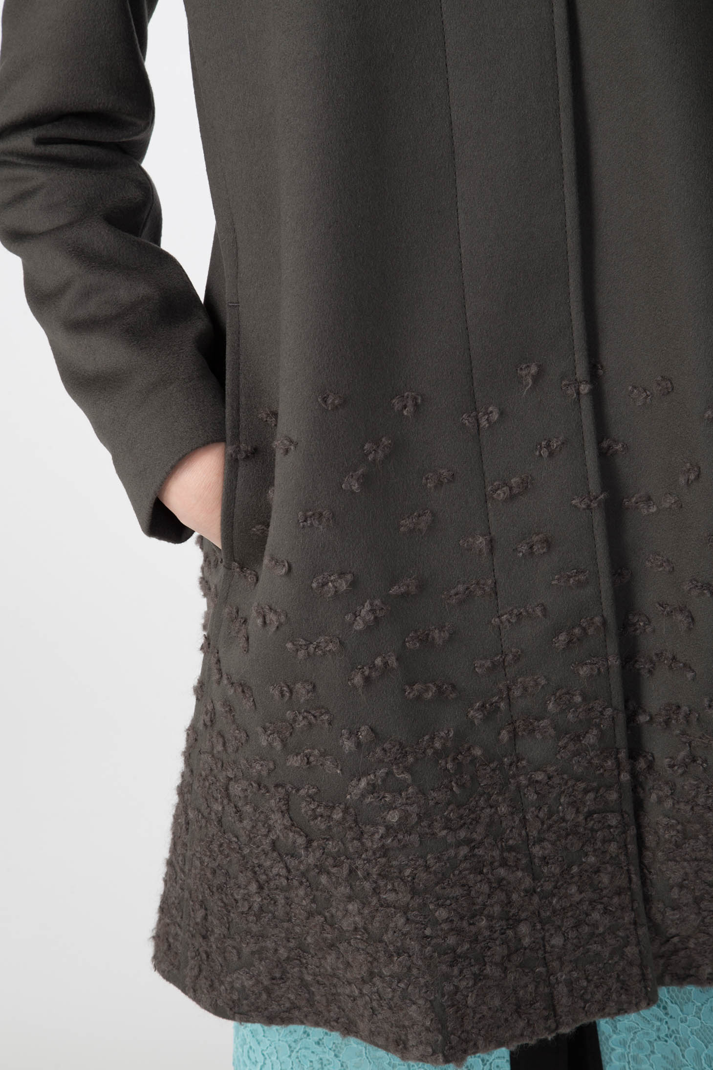 A person wearing the Antonio Croce Carolina Textured Coat with Hood in dark green, featuring a detachable fur hood. Their left hand rests in the pocket of the coat. Light blue lace details are visible beneath the coat, creating a chic winter ensemble against a plain white background.