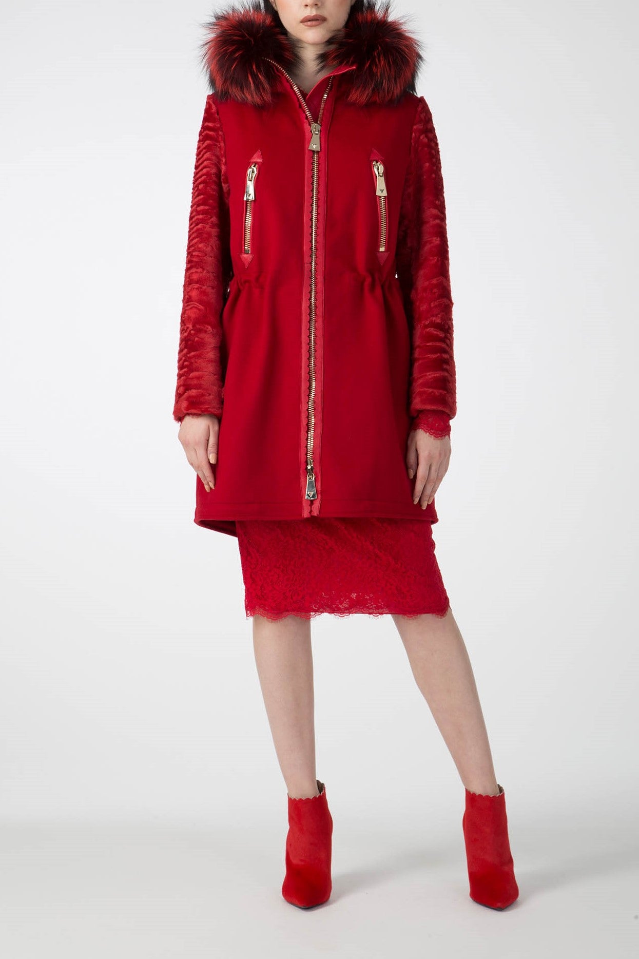 A person is wearing the Antonio Croce Catrina Coat, featuring a vibrant red hue with a luxurious fur collar trim on the hood and elegant ribbed sleeves. The coat’s silver zippers perfectly complement a red lace skirt and ankle boots. Set against the plain white background, this vintage-inspired piece by Antonio Croce truly stands out.