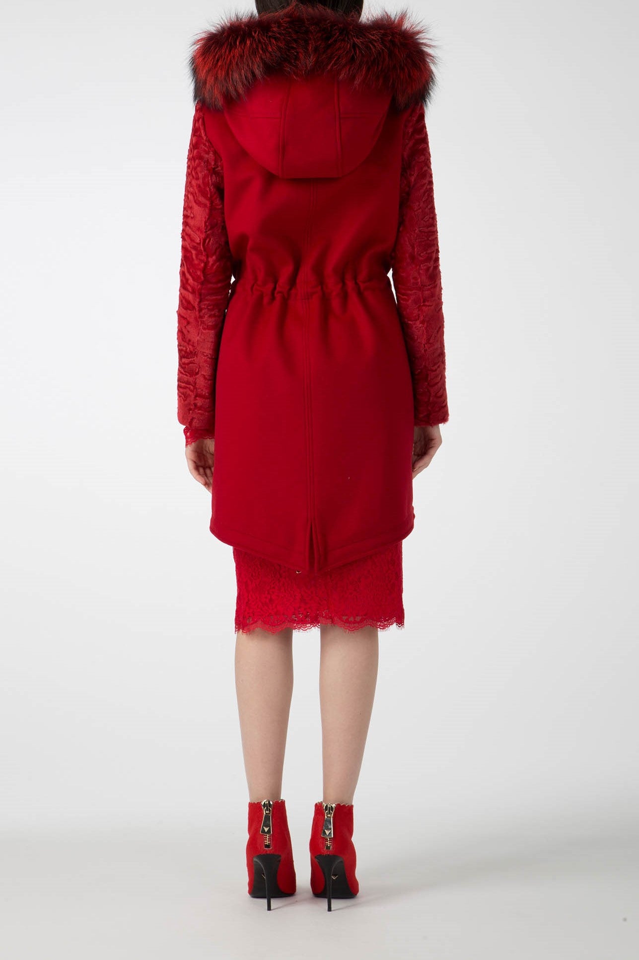 A person is wearing the Antonio Croce Catrina Coat with a fur-lined hood and textured sleeves, standing facing away. This bold red vintage piece is paired with a matching lace skirt and high-heeled ankle boots against a plain white backdrop.