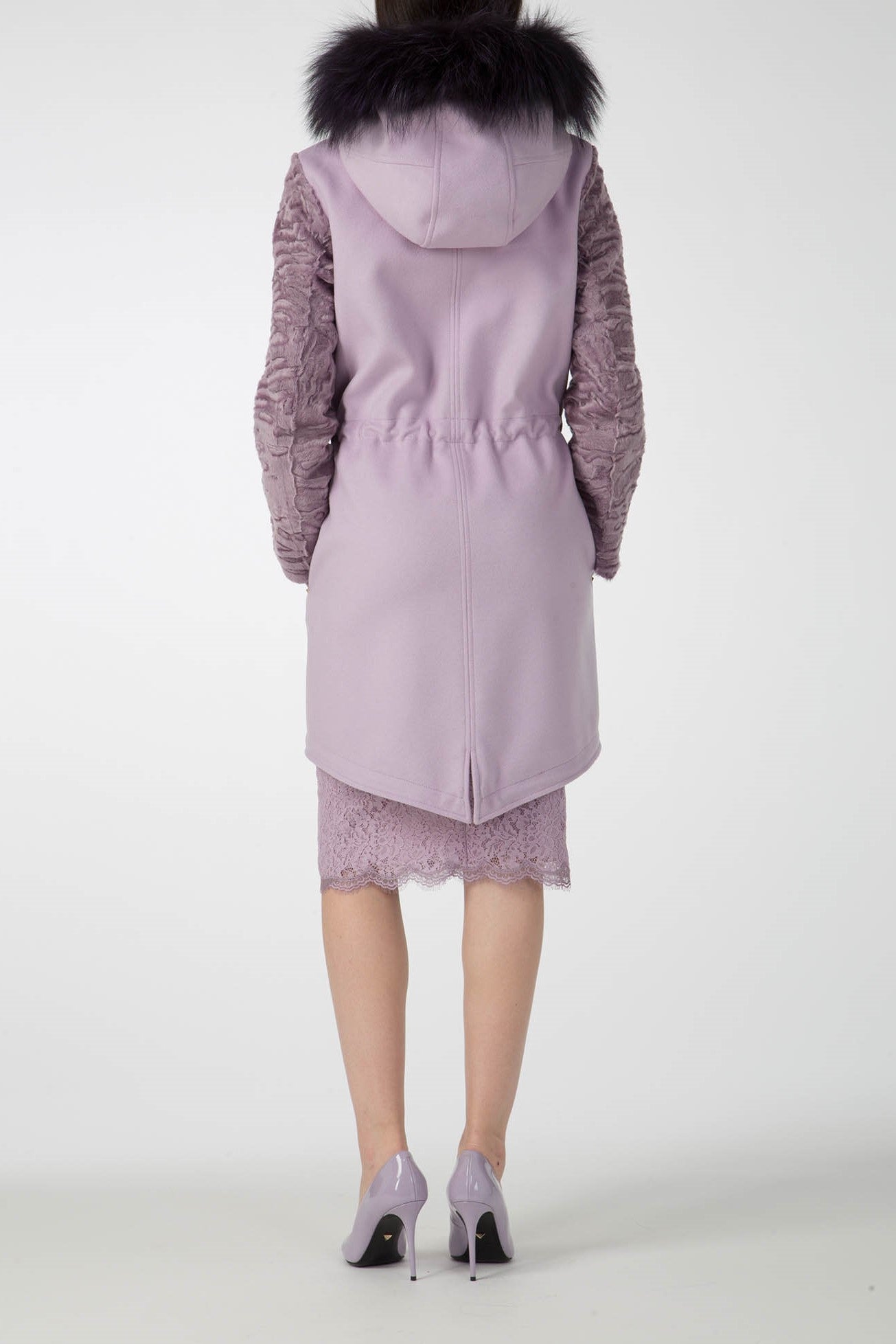 A person is wearing the striking Antonio Croce Catrina Coat, characterized by its bold lavender hue with a fur-trimmed hood and textured sleeves, featuring an asymmetric hem at the back. Complemented by matching lavender high heels and a lace skirt, they stand out dramatically against a plain background.