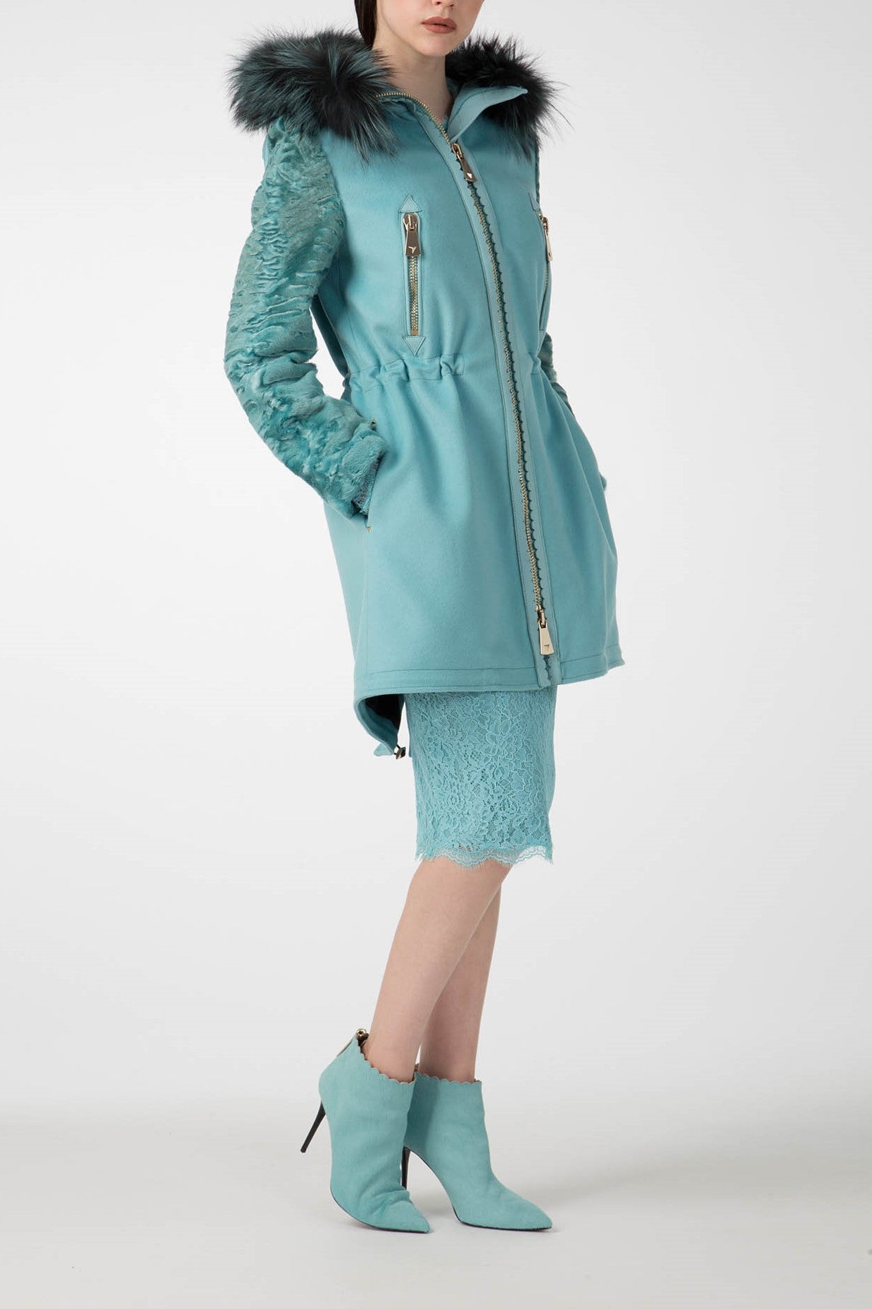 A person is wearing the Antonio Croce Catrina Coat, featuring a bold teal color with a fur-trimmed hood and textured sleeves. They're standing with hands in pockets, paired with a matching teal skirt and ankle boots, creating a striking vintage look against a plain white background.