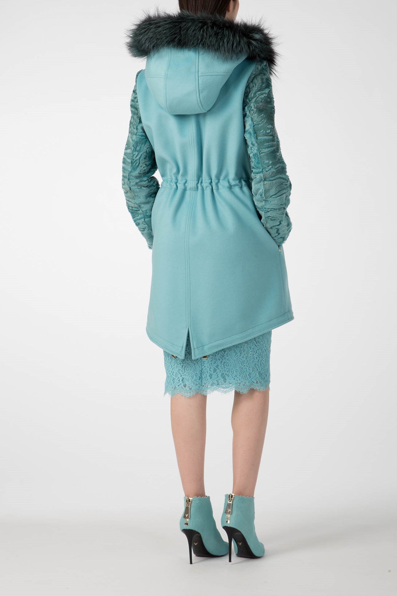 A person wearing the Antonio Croce Catrina Coat with Fur Trim and Textured Sleeves in a bold teal color stands with their back to the camera. They are also dressed in a vintage lace skirt and matching teal ankle boots with gold zippers. The background is white.