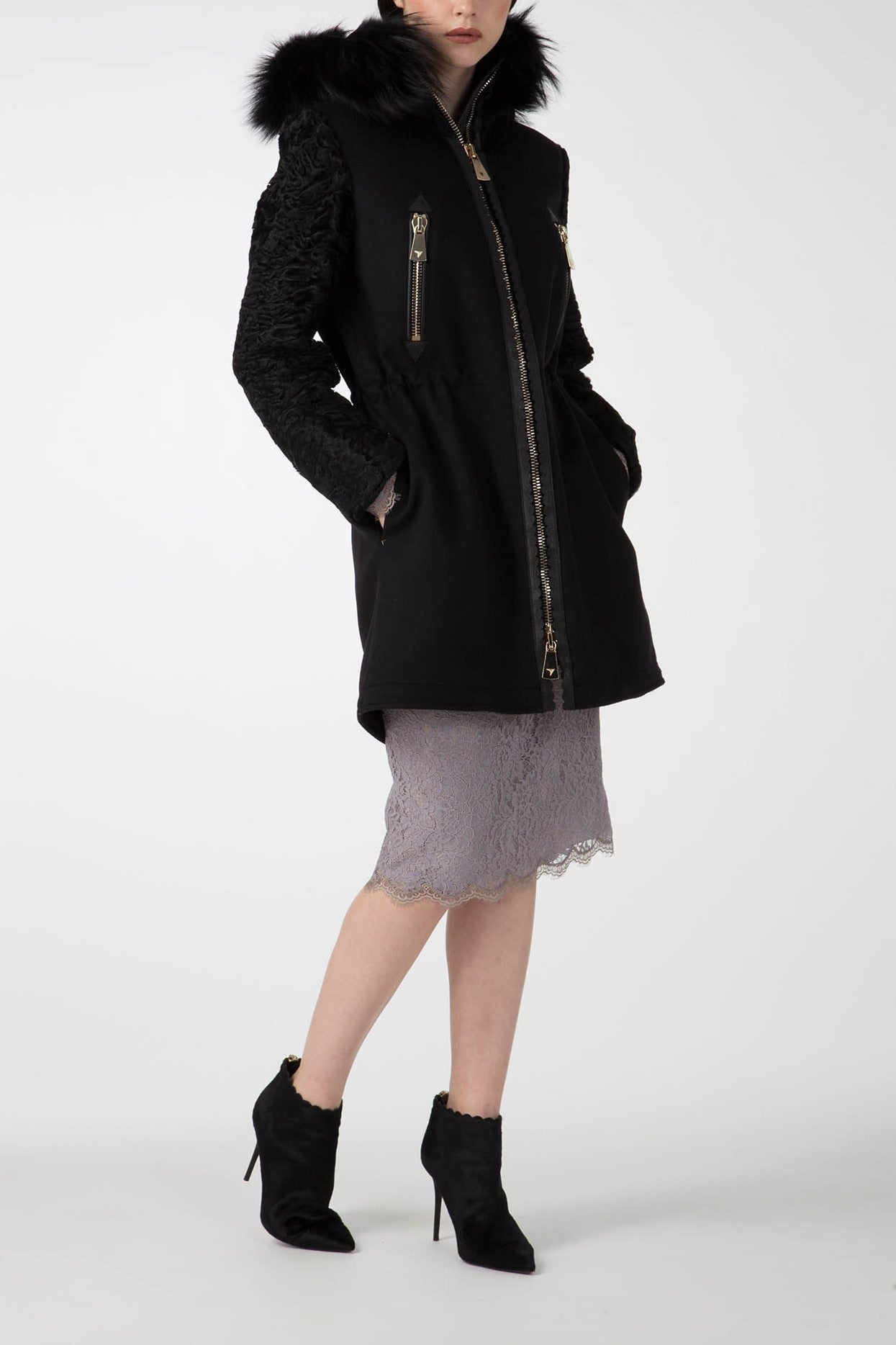 A person is wearing the eye-catching Antonio Croce Catrina Coat featuring a luxurious fur-trimmed collar and textured sleeves, complemented by a lacey gray skirt and chic black ankle boots. The coat's gold zipper details stand out elegantly against the plain white background.