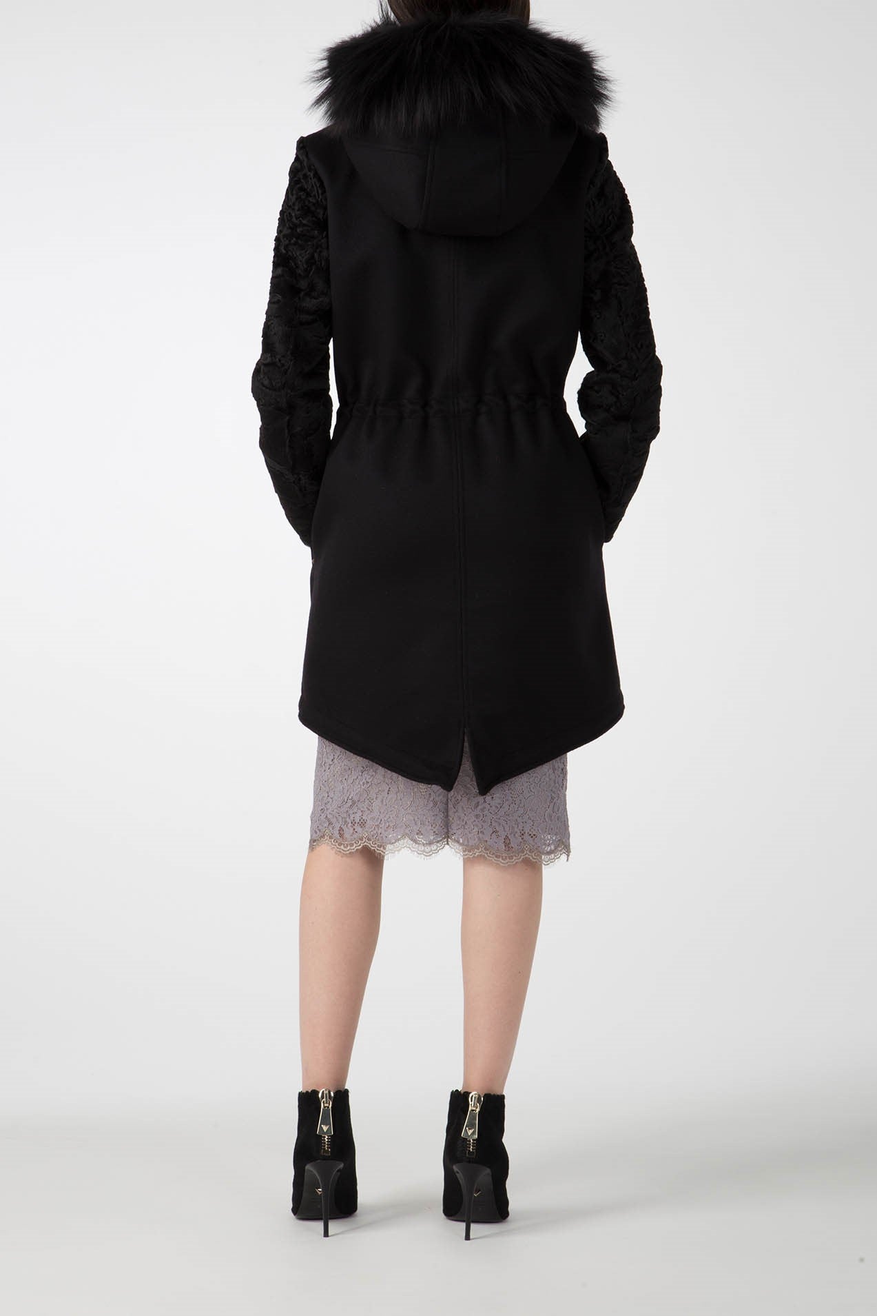 A person stands with their back turned, showcasing the Antonio Croce Catrina Coat, which features a fur-trimmed hood and textured sleeves. This bold piece is paired with an asymmetrical hem over a knee-length gray lace skirt. The look is completed by black high-heeled boots, creating a vintage aesthetic against a plain backdrop.