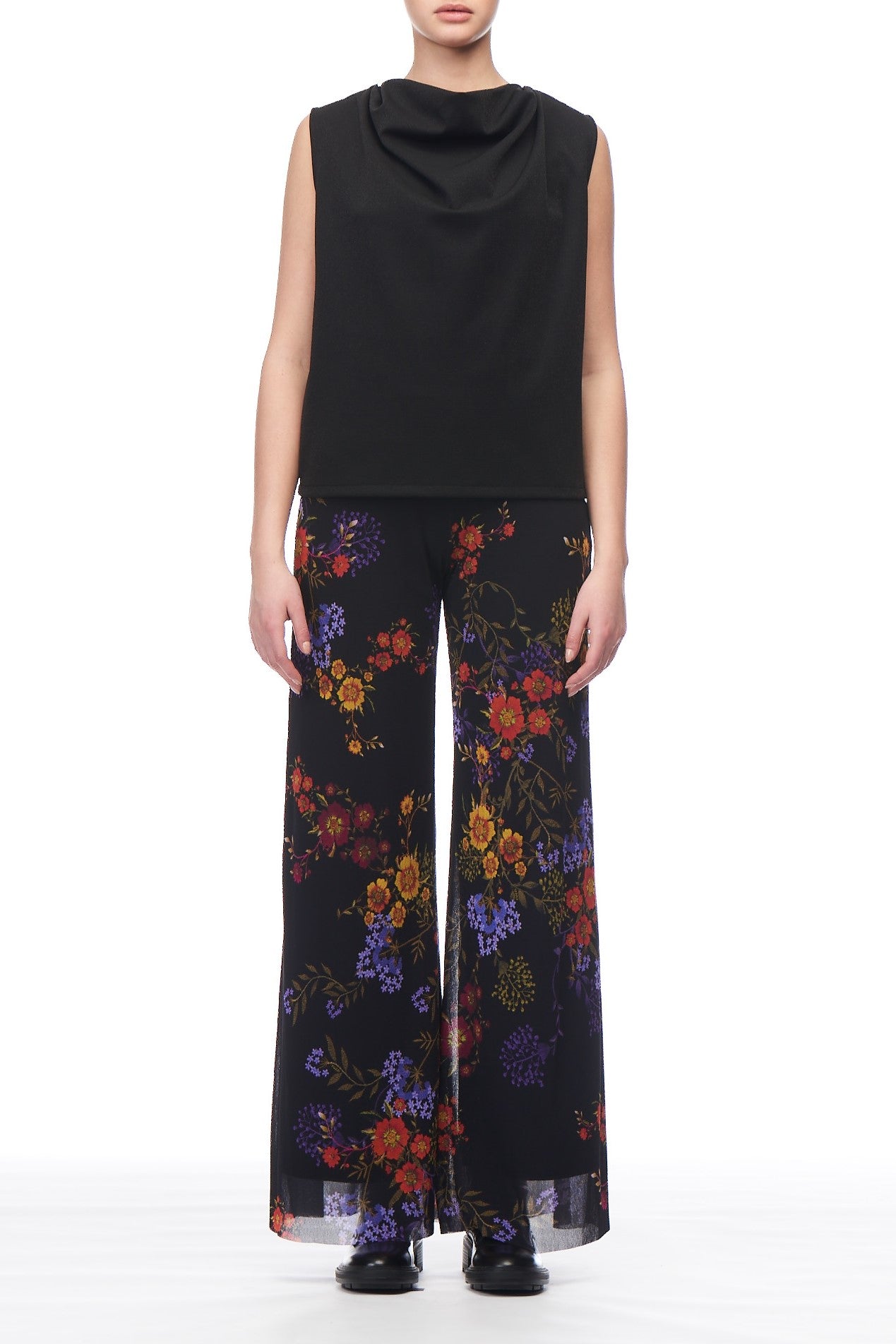 A person in a Fuzzi Woman Cowl Neck Sleeveless Top and wide-leg black pants with colorful floral patterns exudes a modern, stylish vibe against the white background. The ensemble, paired with sleek black shoes, subtly hints at vintage piece inspiration while maintaining its contemporary charm.