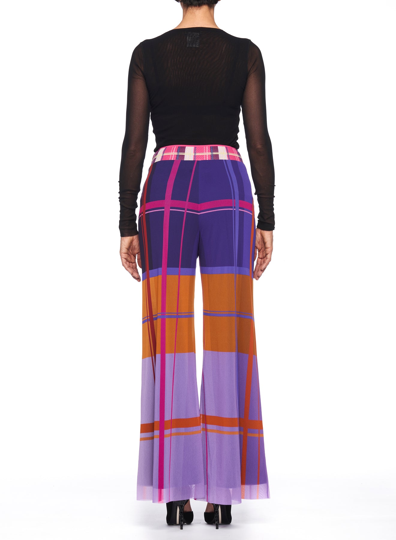 A person stands facing away, showcasing the Fuzzi Woman Colorful Wide-Leg Pants by Fuzzi, which feature a high-waisted fit in vibrant plaid shades of purple, pink, and orange. They pair the pants with a sheer black top and sleek black high-heeled shoes against a simple white backdrop.