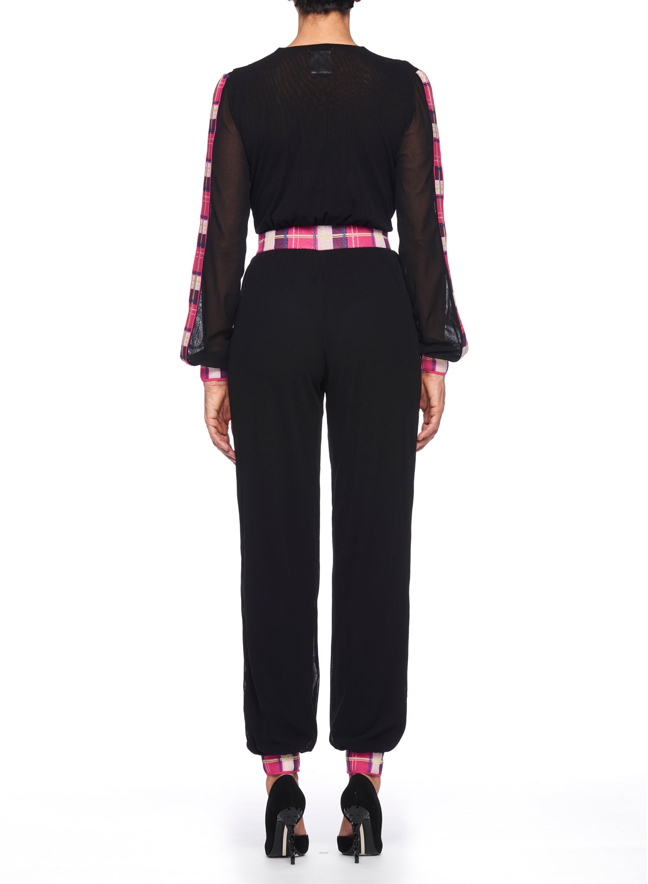 A rear view of a person wearing the Fuzzi Woman Black Mesh Jumpsuit with Colorful Details, featuring sheer sleeves and pink plaid accents. The outfit is completed with long pants and high heels, set against a crisp white background.