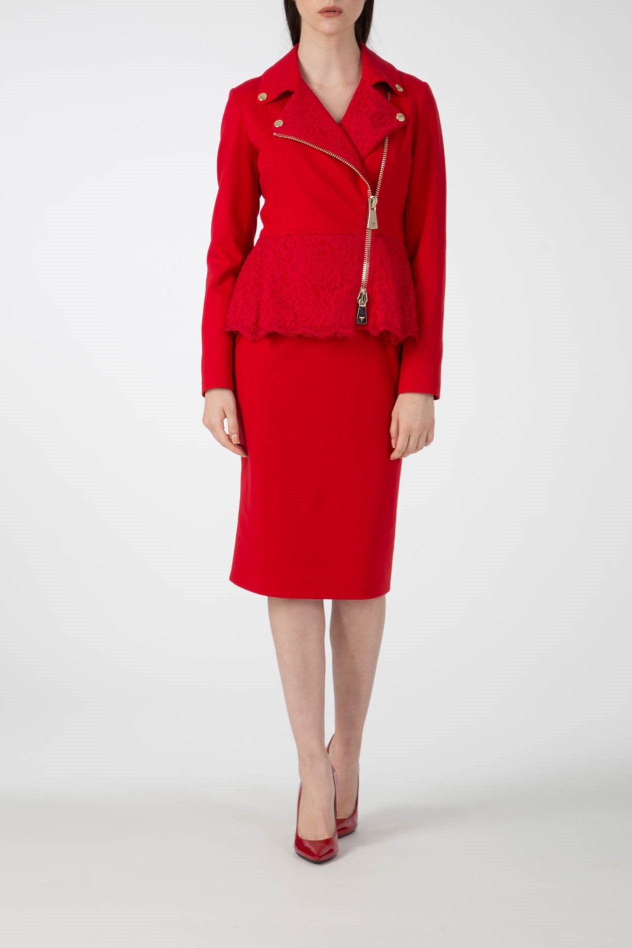 A person is wearing a chic red ensemble featuring the Antonio Croce Gigi Jacket with gold hardware, characterized by its edgy design and asymmetric zipper, complemented by a matching pencil skirt. The look is completed with red high-heeled shoes, all set against a plain white background.