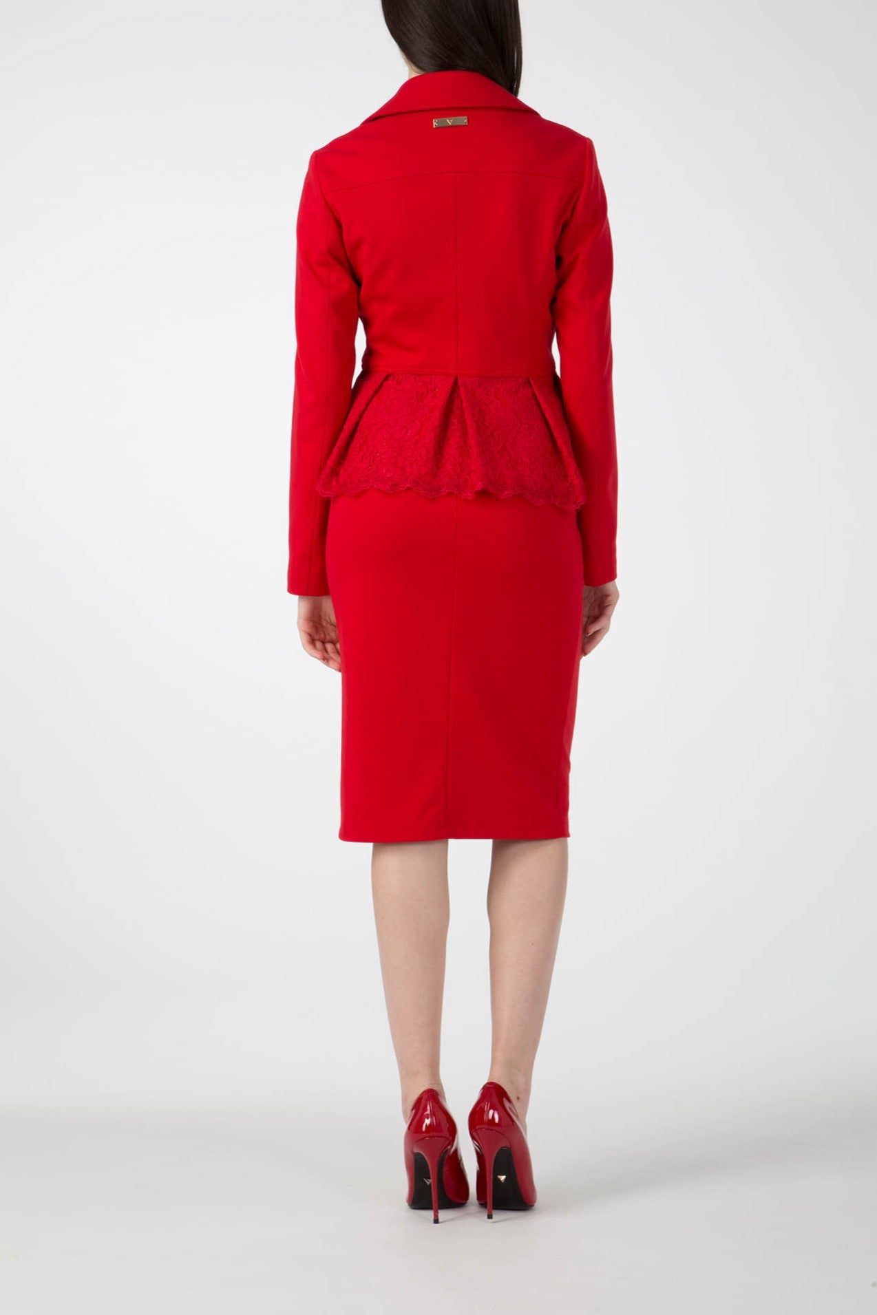 A woman in an Antonio Croce Gigi Jacket with gold hardware, fitted and featuring an edgy peplum design with an asymmetric zipper, stands facing away. She pairs it with matching red high heels against a plain white background.