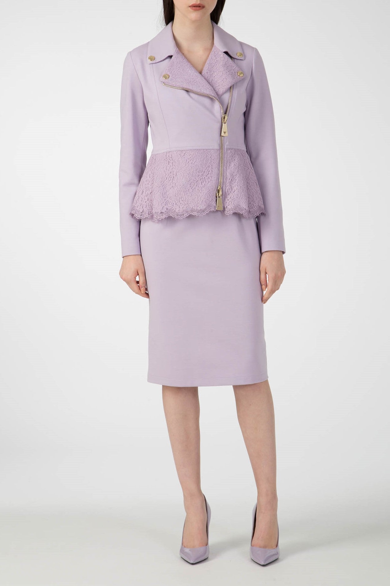 A woman is dressed in a light purple dress, complemented by the Antonio Croce Gigi Jacket, which stands out with its edgy peplum style, asymmetric zipper, and gold hardware. She completes her outfit with purple high heels, all set against a plain white background.