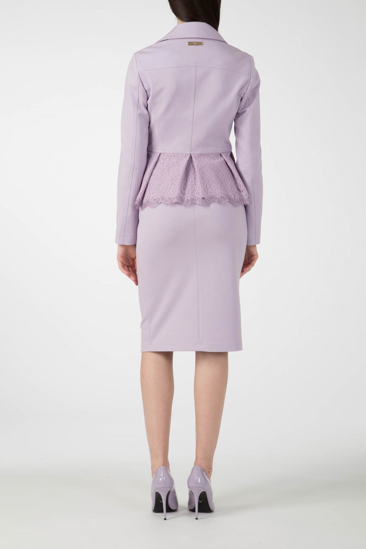 A person stands facing away, wearing the Antonio Croce Gigi Jacket with gold hardware in pastel lavender, featuring an edgy peplum detail and an asymmetric zipper. The outfit is perfectly matched with a pencil skirt of the same hue and lavender high-heeled shoes against a plain white background.