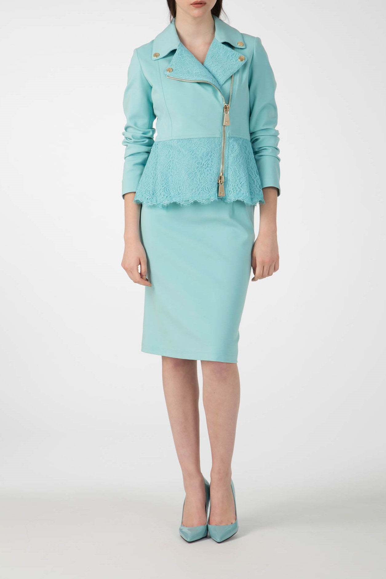 A woman is wearing a teal suit comprising the Antonio Croce Gigi Jacket with Gold Hardware and a pencil skirt. The jacket, known for its edgy design, features an asymmetric gold zipper and decorative buttons. She completes her stylish ensemble with matching teal heels while standing confidently against a plain white background.