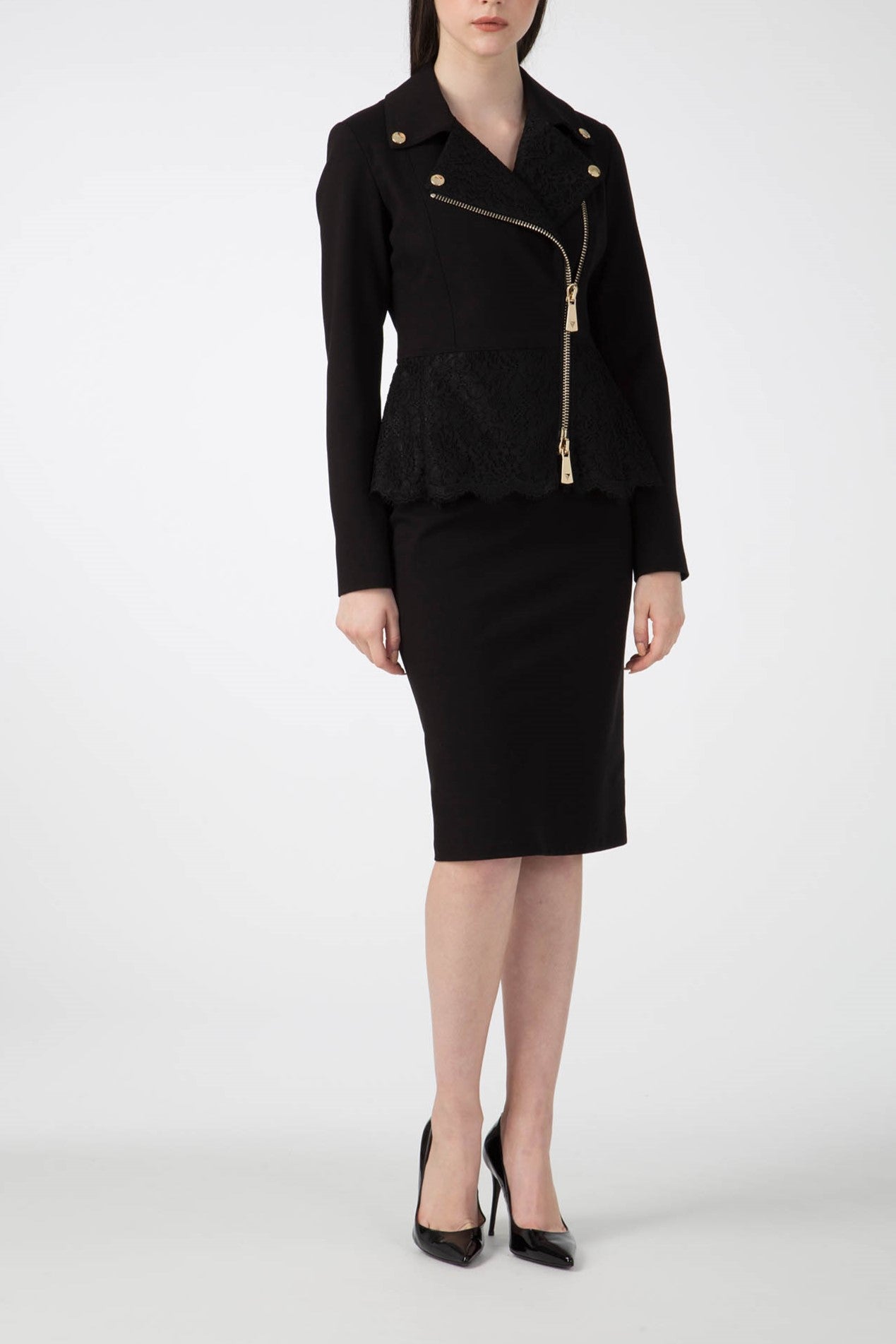 A woman in the edgy Antonio Croce Gigi Jacket with gold hardware and an asymmetric zipper, paired with a black lace peplum top and knee-length pencil skirt. She is wearing black high heels, standing against a plain white background. Her face is not visible.