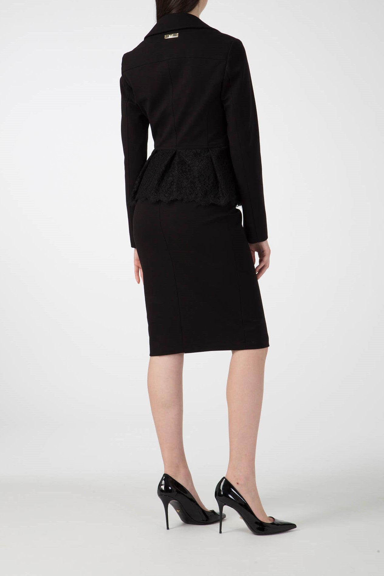 A person facing away, wearing an Antonio Croce Gigi jacket with gold hardware, styled in a black business suit. The outfit includes a peplum jacket with an asymmetric zipper paired with a pencil skirt and complemented by black high heels, all set against a plain white background.