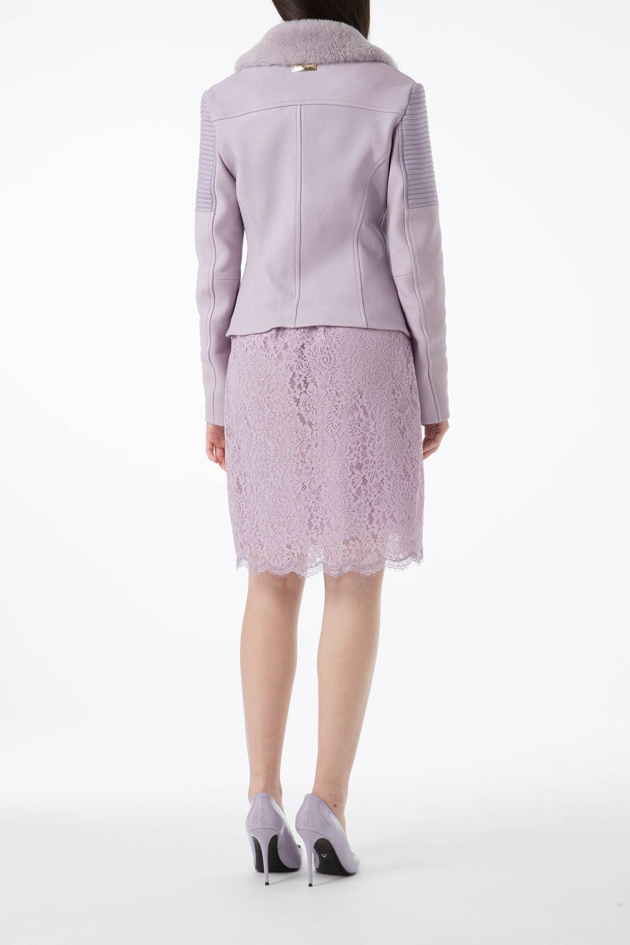 A woman in a light purple lace dress with an edgy vibe stands against a plain white background, wearing the Antonio Croce Green Leather Jacket with Fur Collar, featuring an asymmetric zipper closure, and high heels with her back to the camera.