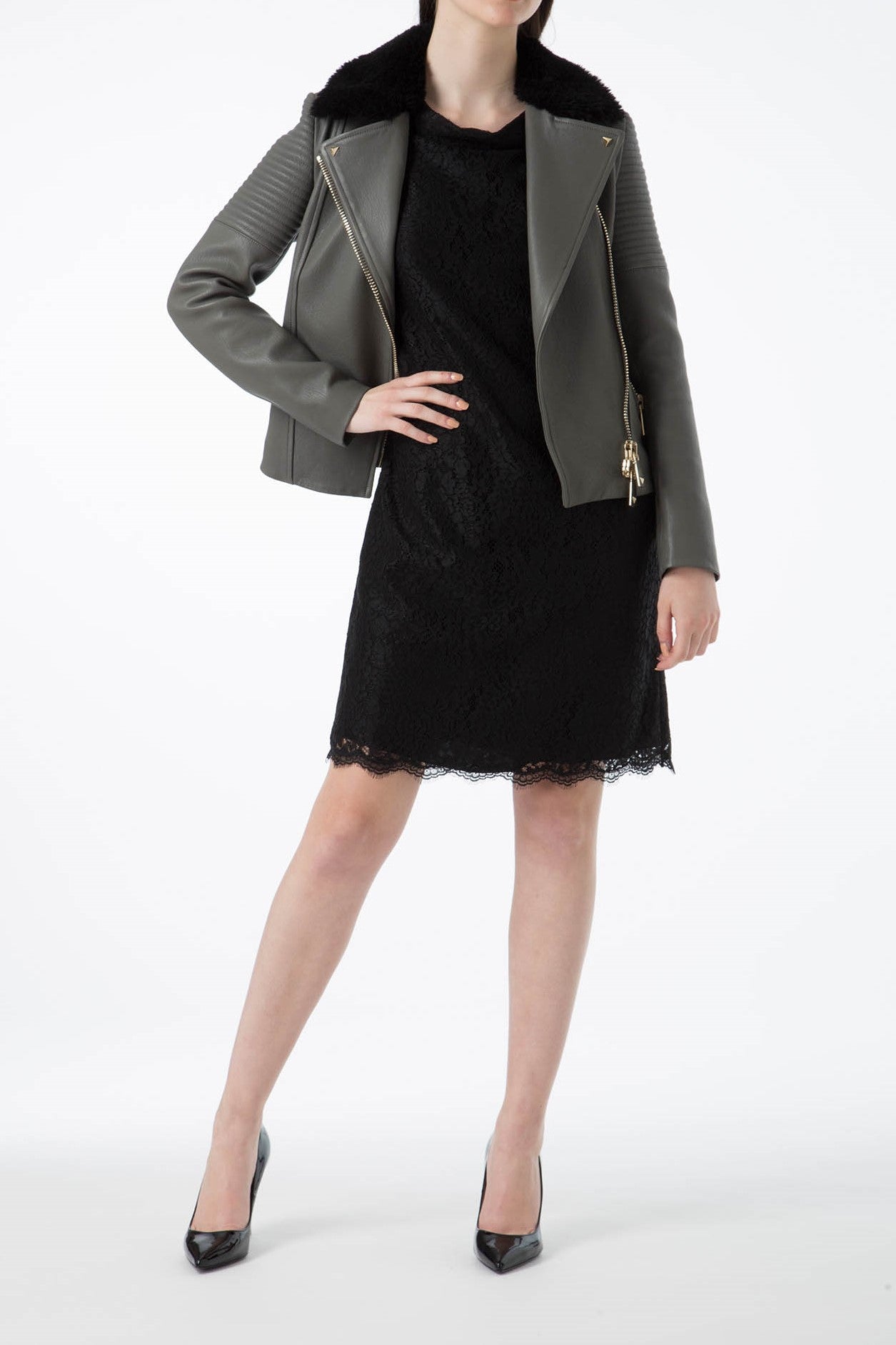 A person in a green leather jacket with a fur collar from Antonio Croce, featuring an asymmetric zipper closure, stands with one hand in a pocket. They are wearing black high heels and a black lace dress against a plain white background.