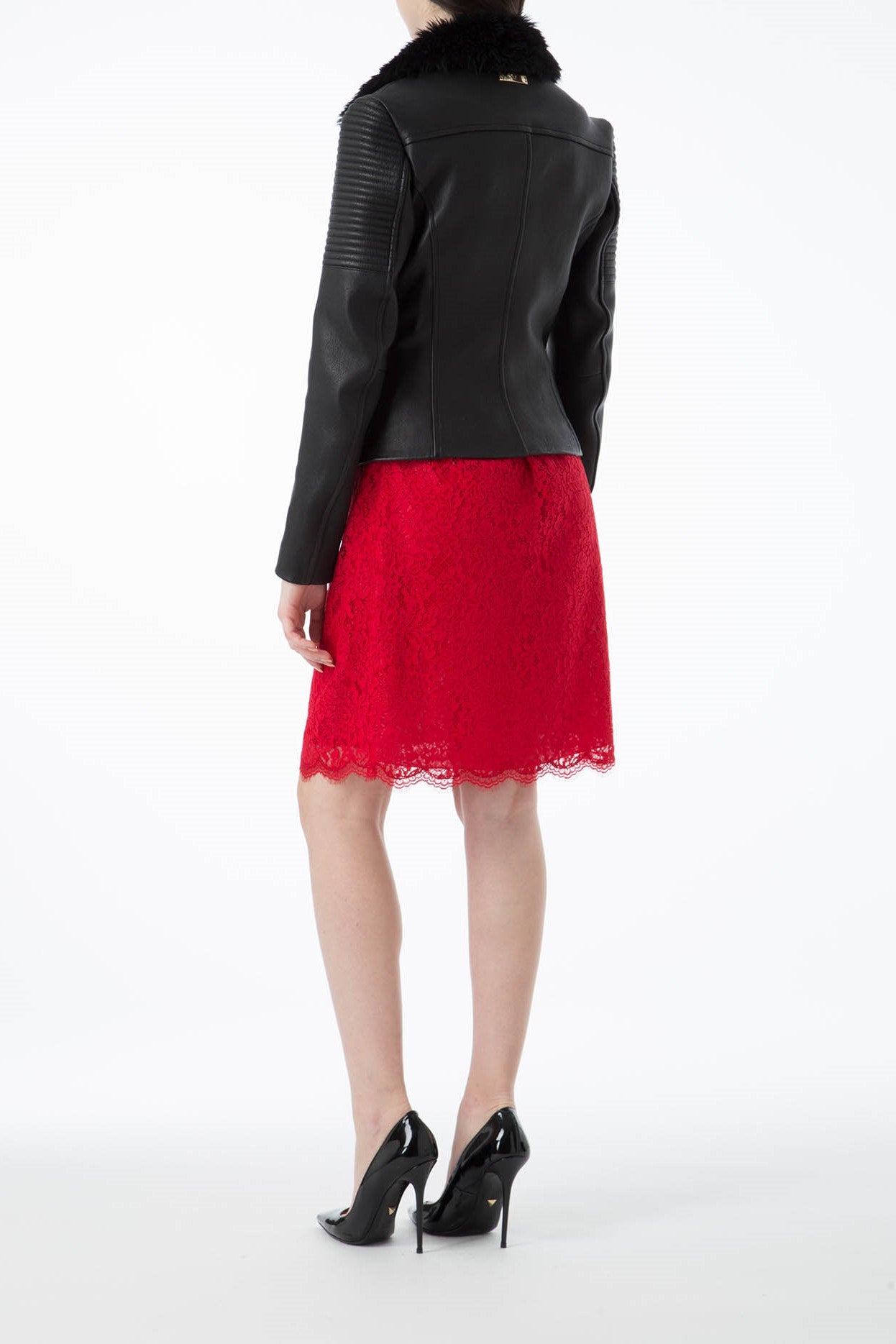 A person in a chic green leather jacket with a fur collar by Antonio Croce and a red lace skirt stands with their back to the camera. They complete the look with black high-heeled shoes against a plain white background.