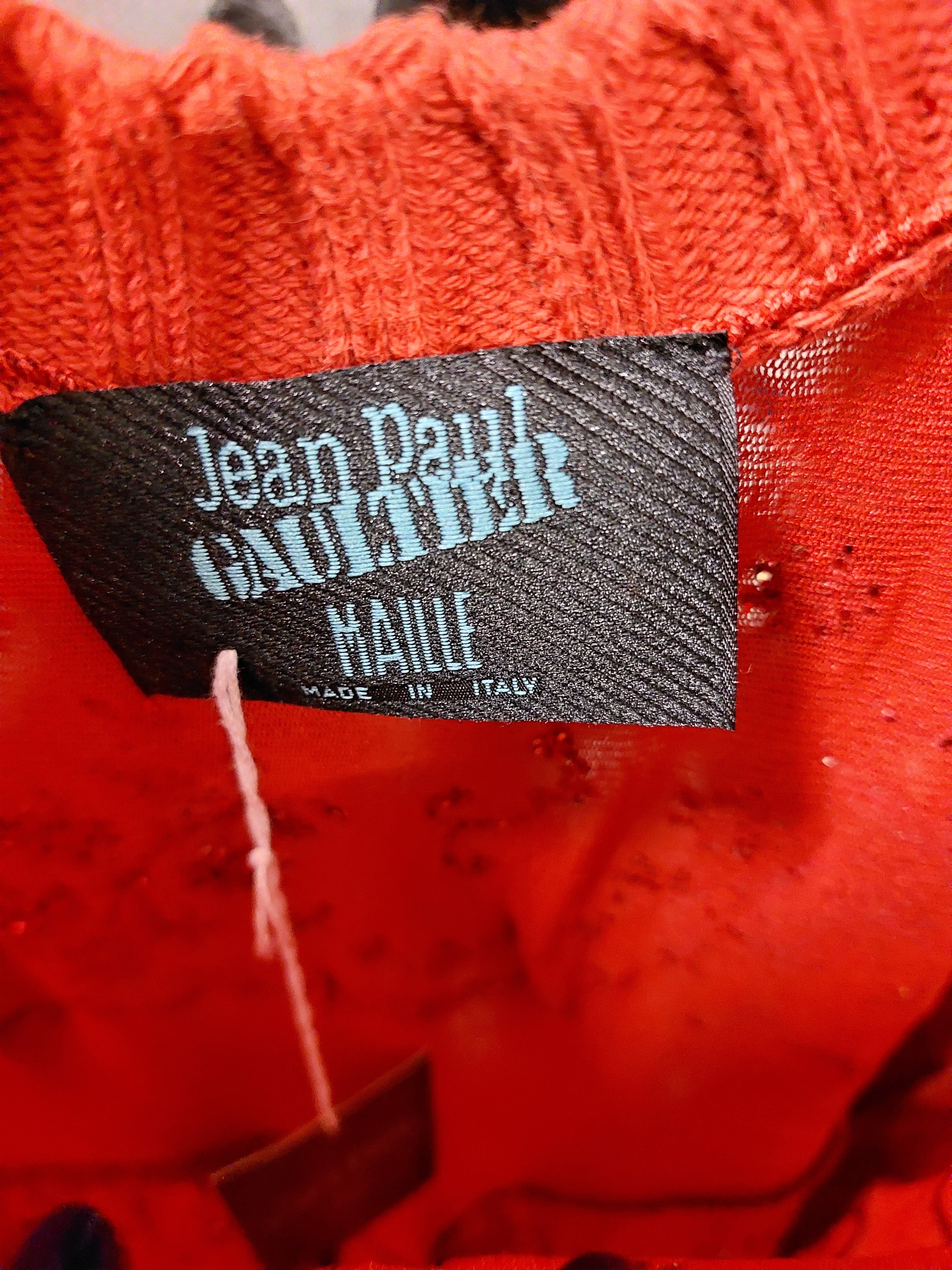 Here's a close-up of a clothing label displaying the brand name JEAN PAUL GAULTIER in blue, alongside "MAILLE MADE IN ITALY" in white, both set against a black background. This label is attached to the Vintage Woman Red Dress from the Spring/Summer 1995 collection, capturing the essence of timeless allure.