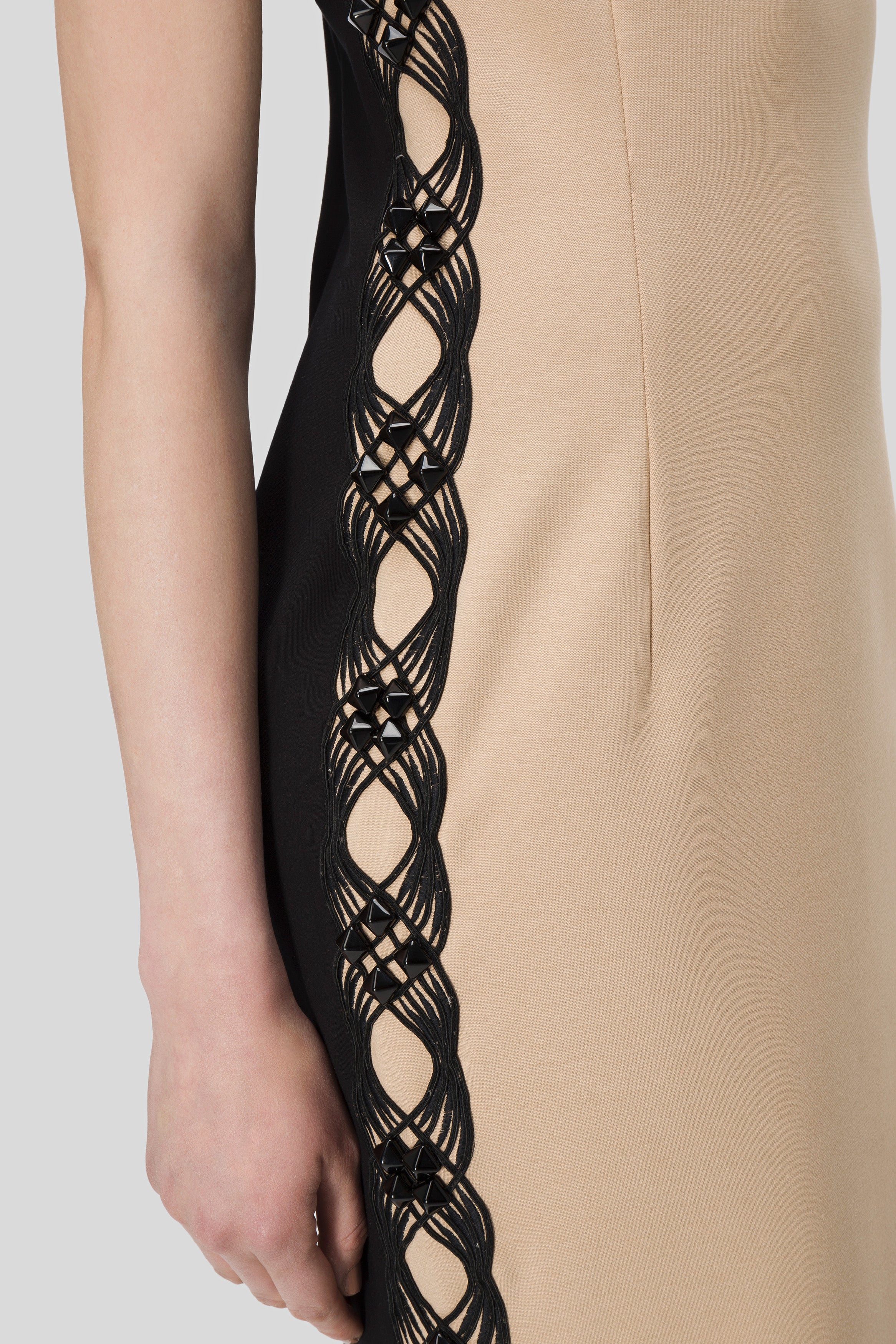 Close-up of an elegant Antonio Croce Adelin Sleeveless Dress featuring a beige front and a striking black side panel. The strapless neckline enhances its sophistication, with the panel showcasing intricate black lattice detailing that subtly reveals the skin beneath. You can see the arm of a person wearing this stunning dress by Antonio Croce.
